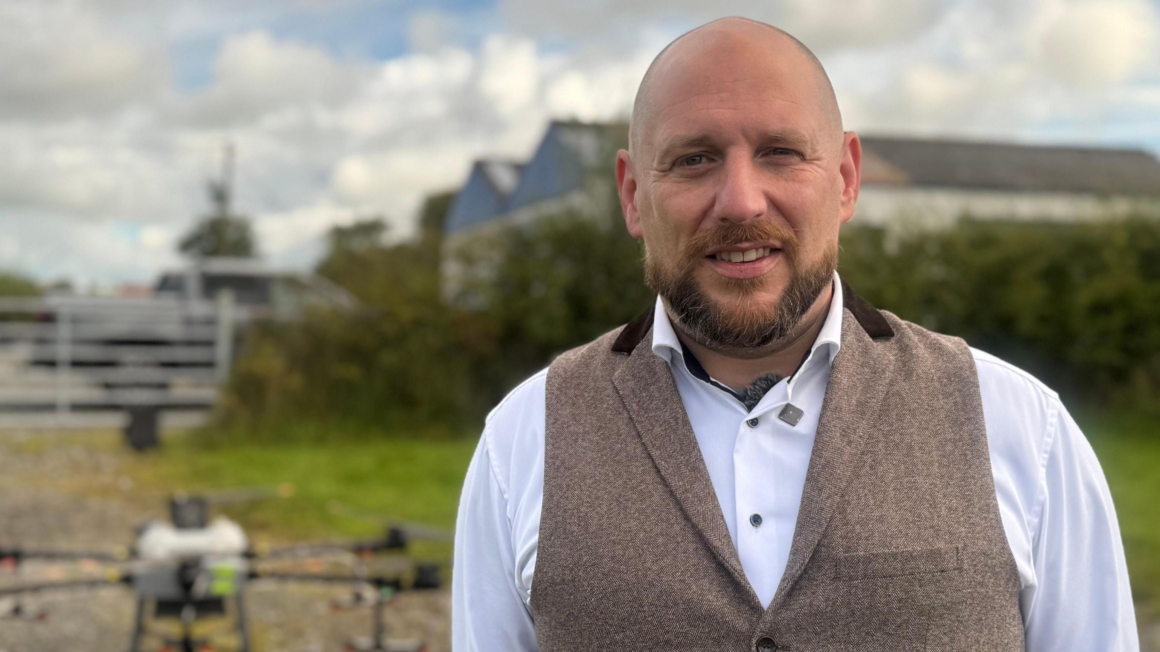Roy Spence standing outside beside the drone, he is wearing a white shirt and a brown waistcoat. He is bald and has a brown beard.