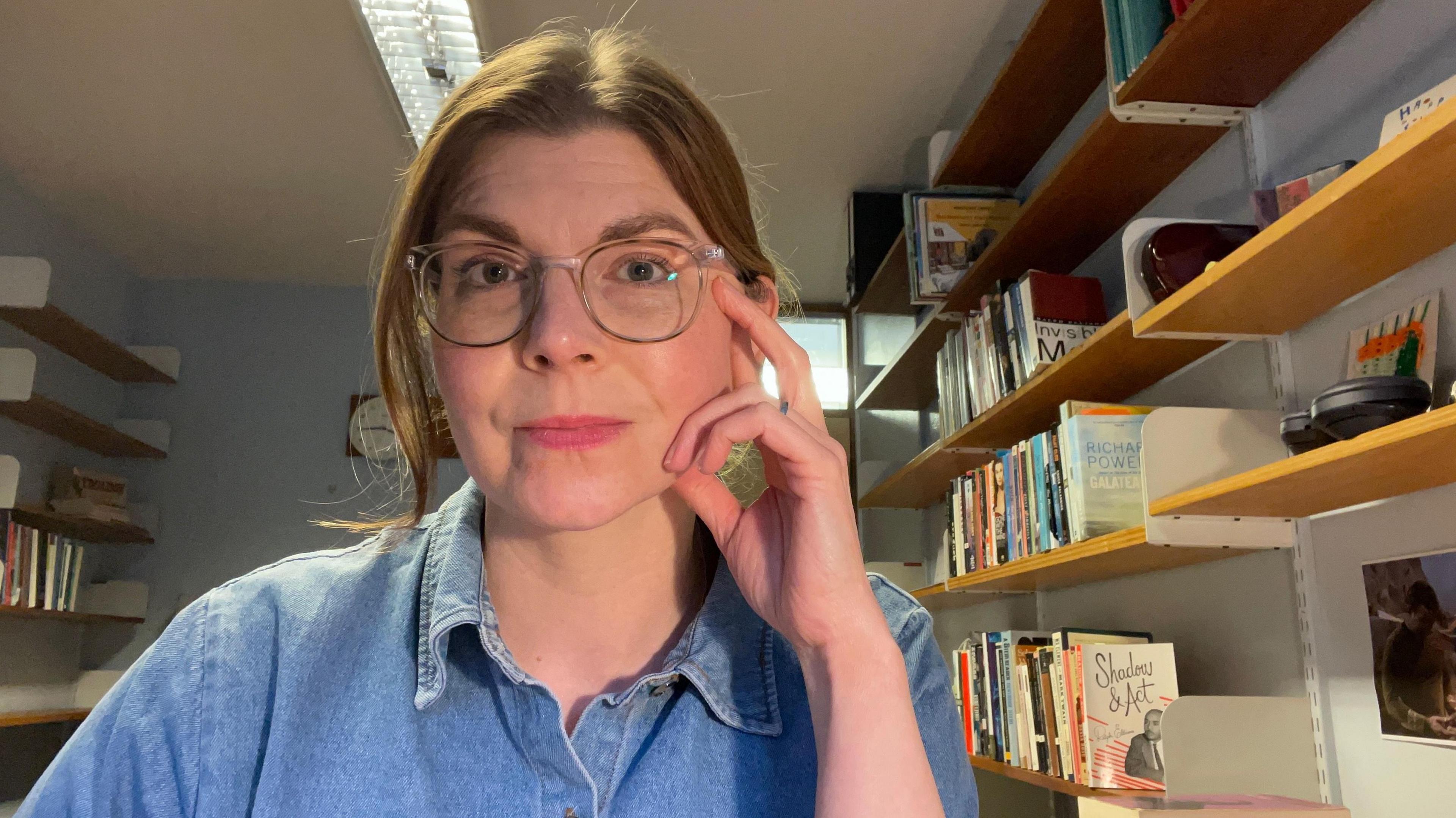 Dr Katie Muth in her office. She is wearing glasses and a blue denim shirt, and has bookshelves behind her.