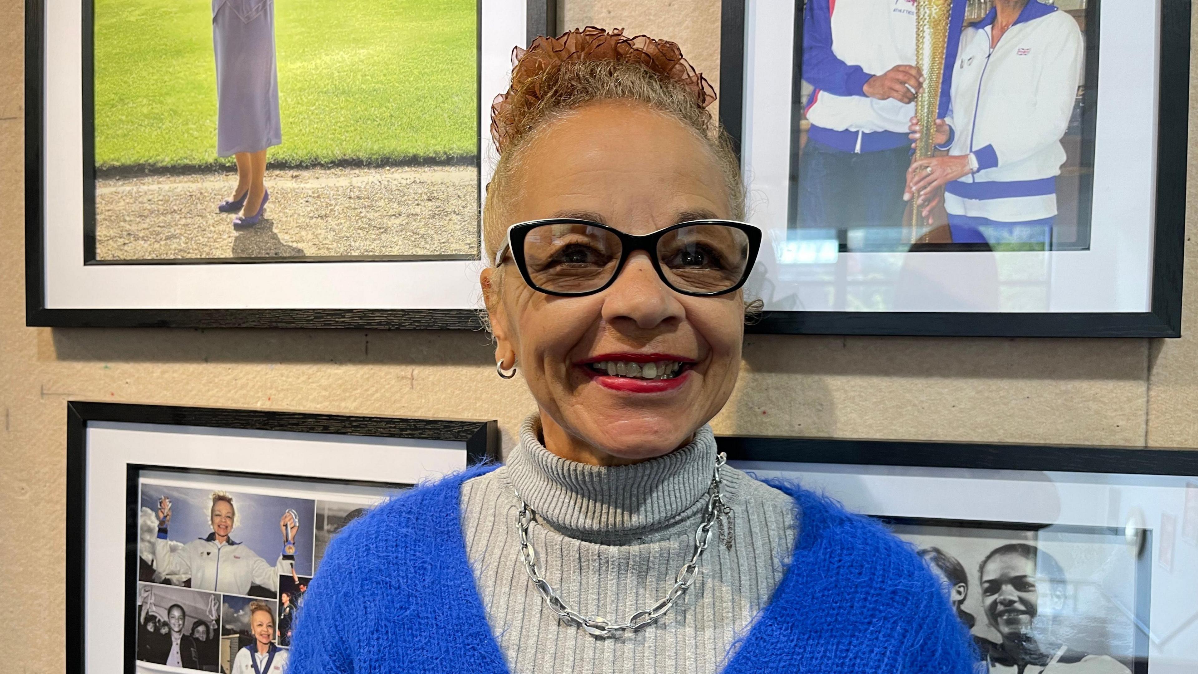 Anita Neil is standing in front of photographs she has contributed to the Black History Month exhibition. She is smiling, has her tied up in a bun, and is wearing black glasses, a grey turtleneck jumper with a blue jumper over the top, and a silver necklace. 