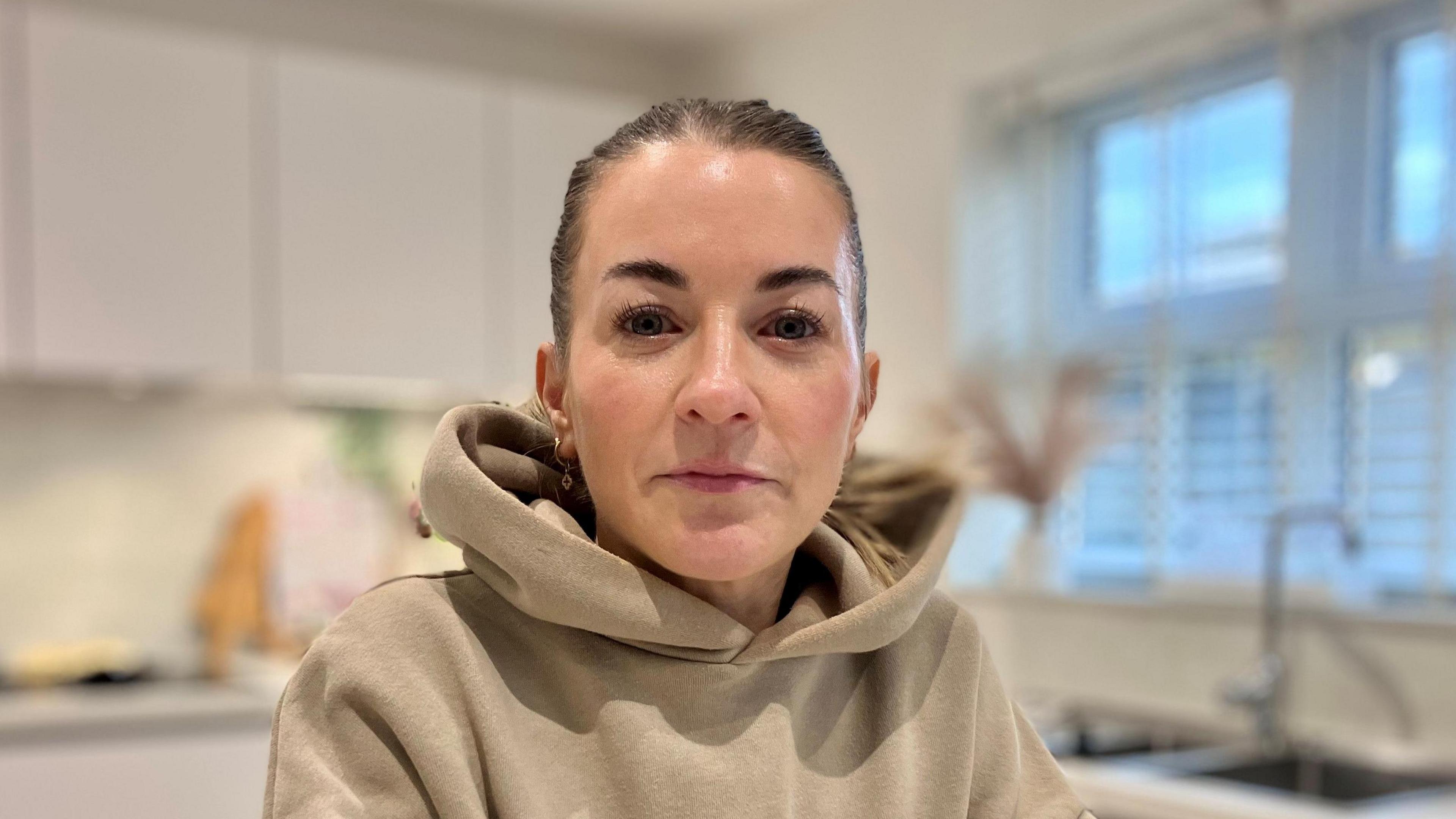 A person wearing an oversized beige hoodie seated at a white countertop in a modern kitchen. The kitchen features white cabinets, a sink under a large window with blinds.