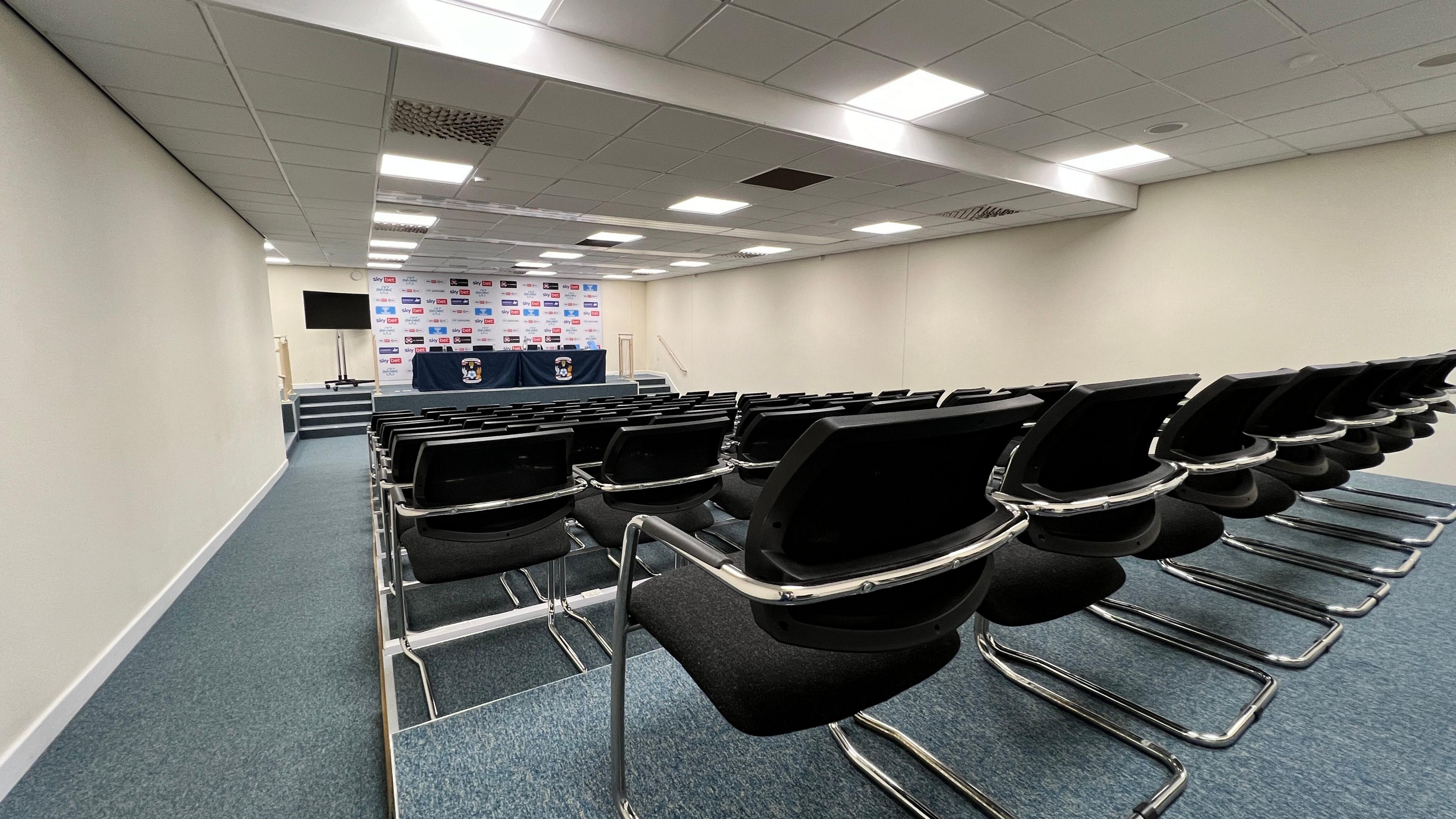 Coventry City's press room, rows of empty chairs are visible and at the back of the room is a long desk