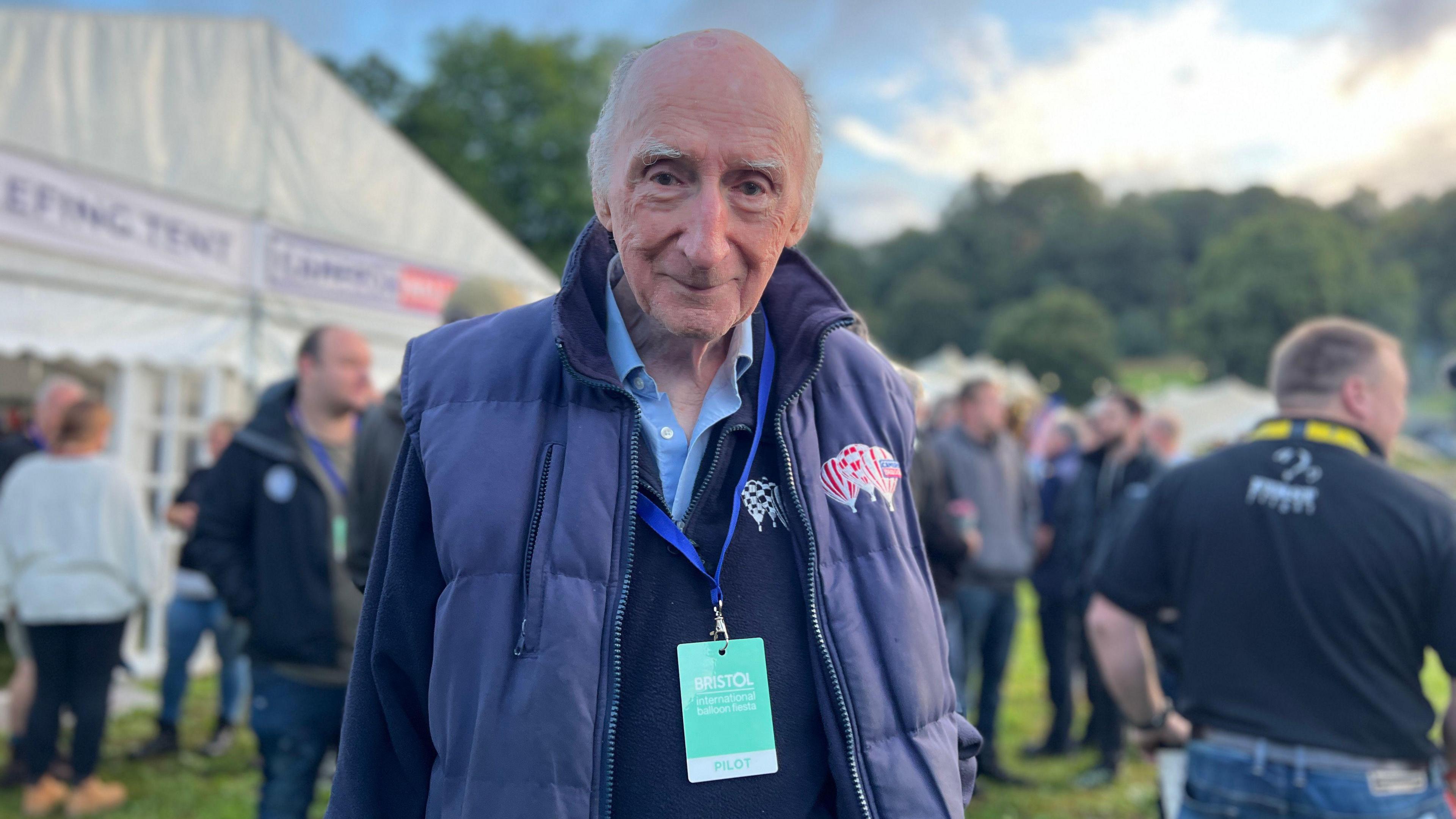 Don Cameron, founder of the fiesta, pictured outside the tent where the briefing takes place. He wears a light blue shirt, navy jumper and blue gilet with hot air balloons embroidered on it. 