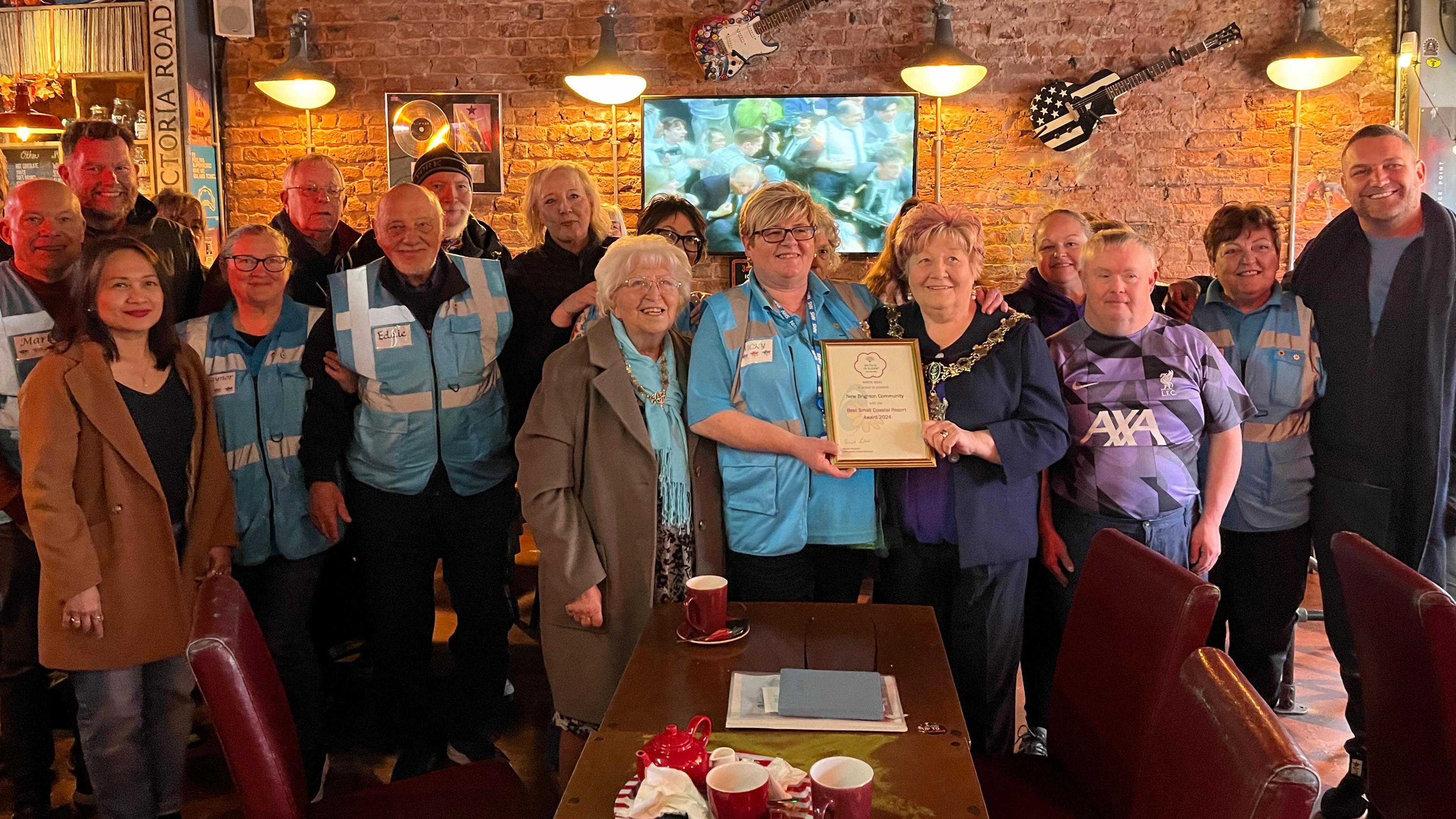 A group of 20 people stand and smile at the camera in a cafe, holding a framed award 