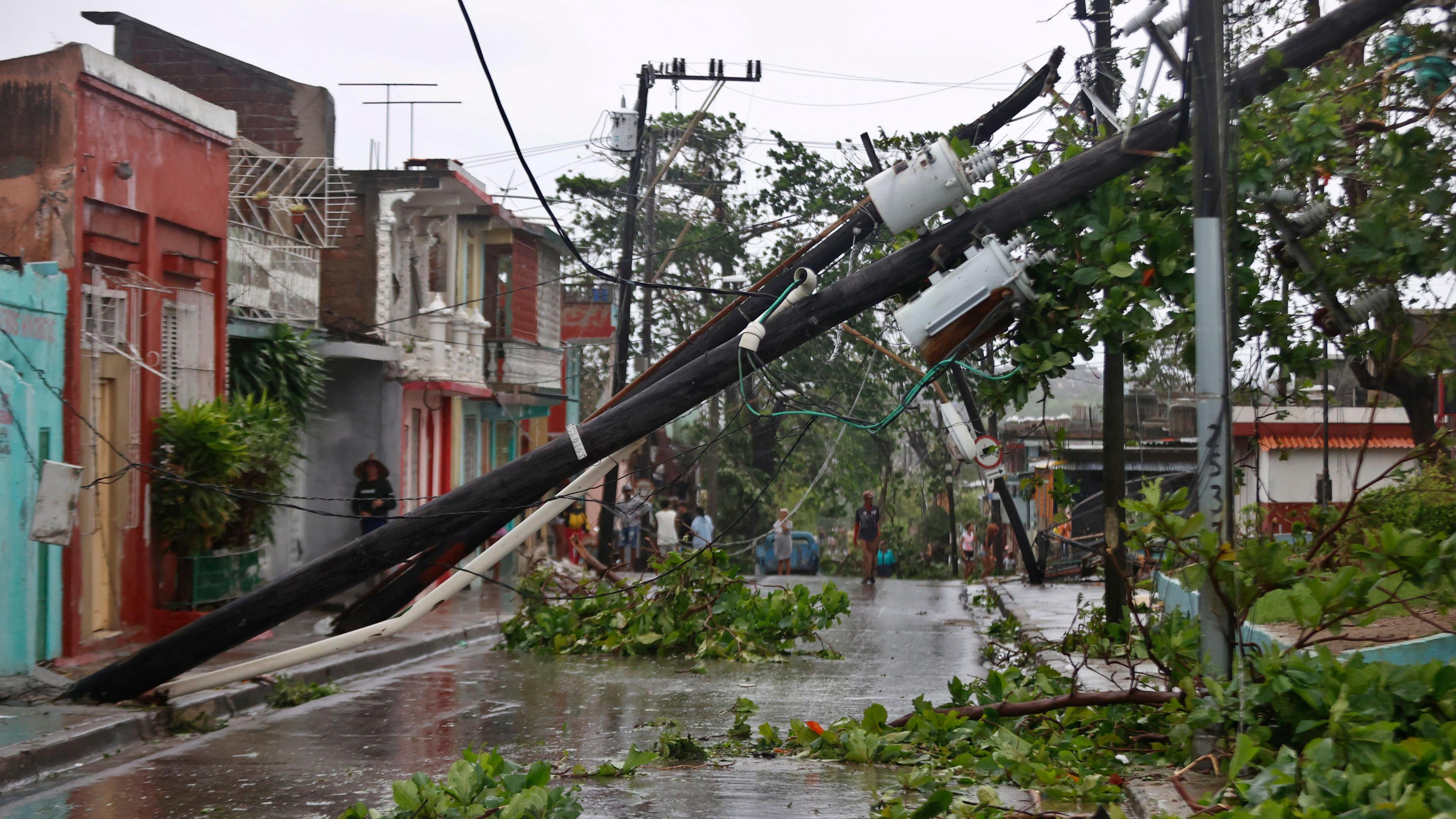 A street affected by Hurricane Melissa in Santiago de Cuba, Cuba. A fallen telephone pole and fallen trees. The ground in wet and there are tree branches on the ground. The sky is grey. There are people walking in the street.