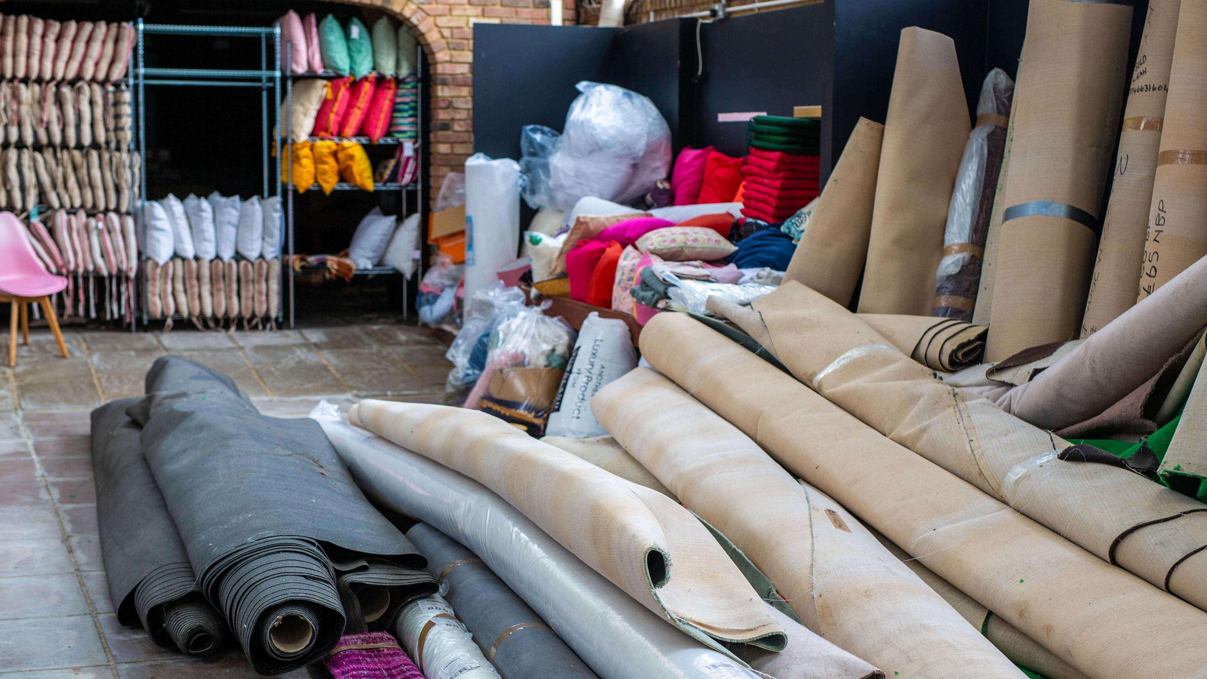 A pile of rolled up carpets on stone tiles. In the background there are shelves with cushions