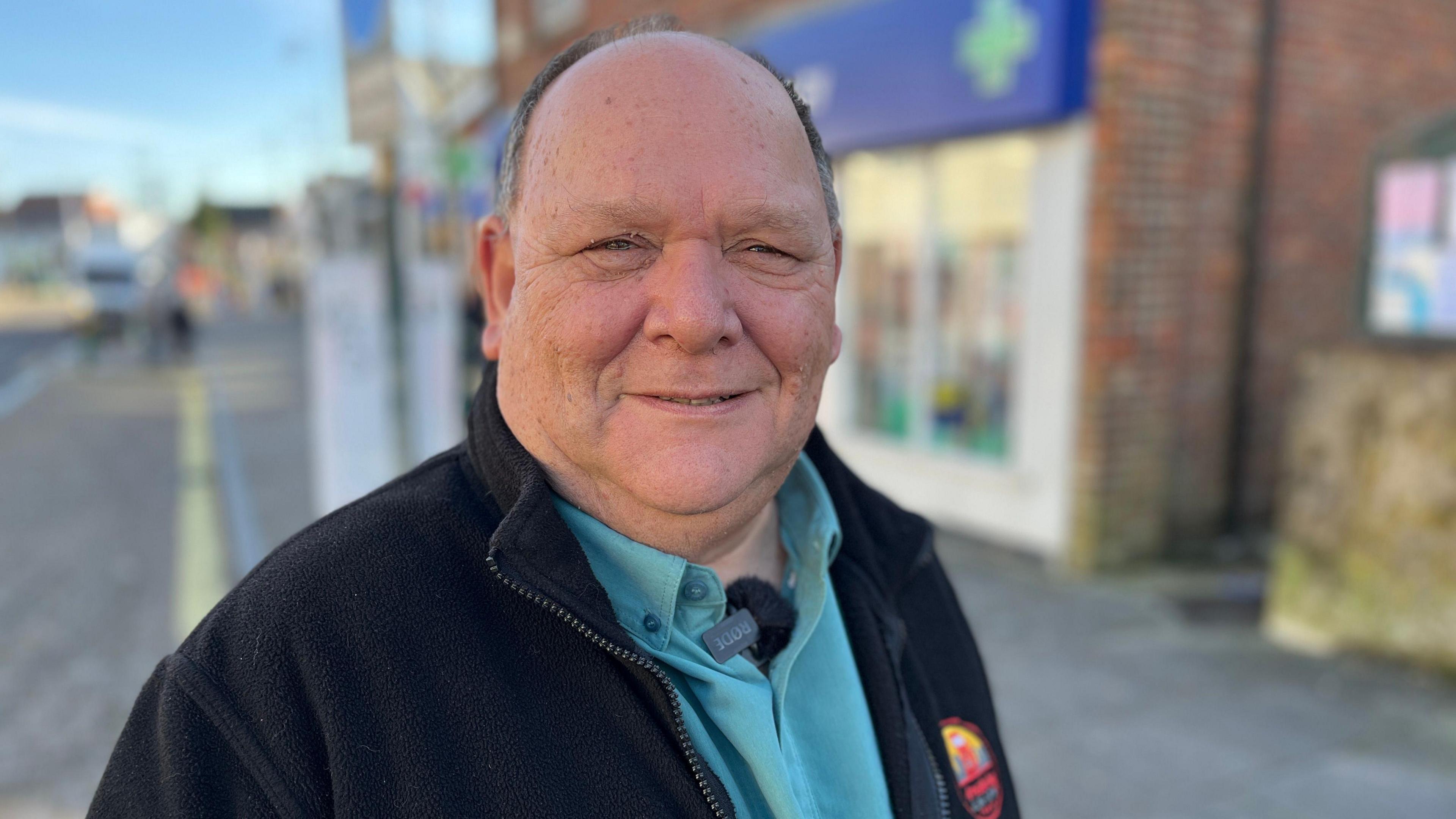 A man wearing a black jacket and blue shirt looks at the camera. The background is blurred, but you can make out a street on the left and a shop with a blue board and a green cross, often used to indicate a pharmacy.