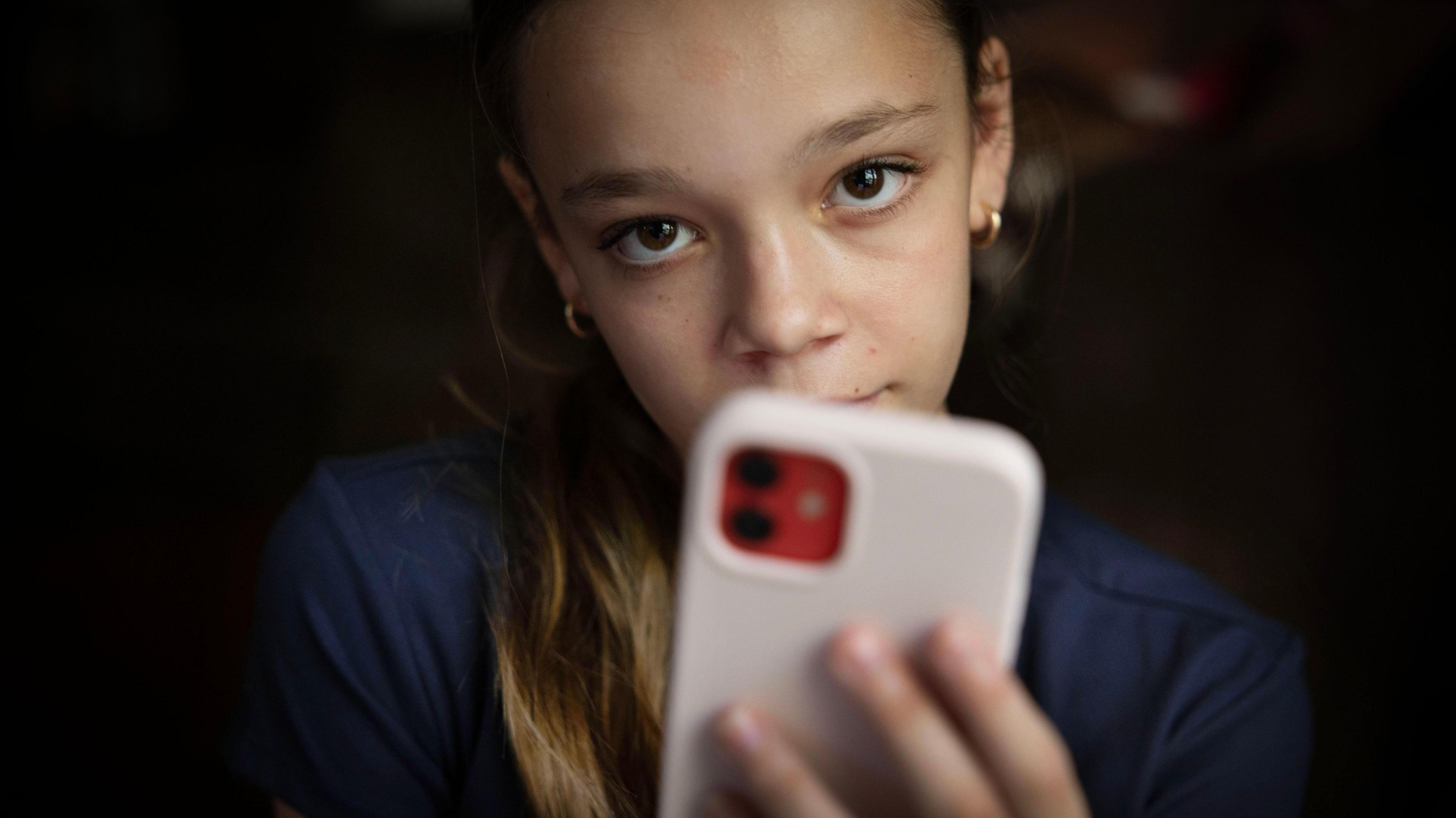 A teen wearing a navy shirt looks at the camera, her pink phone partially covering her face