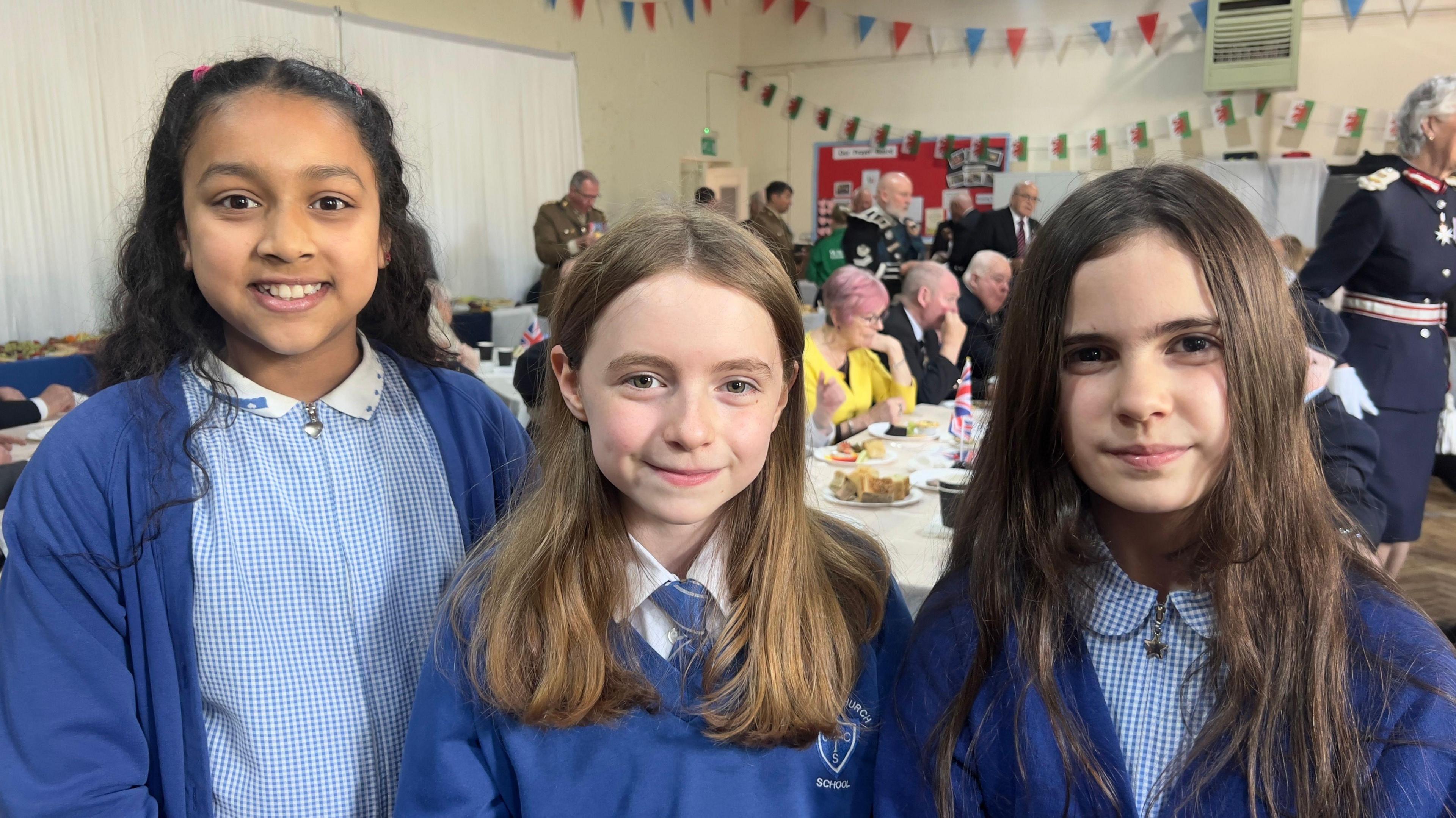 Three young girls wearing blue uniforms. They are smiling at the camera while standing in a classroom.