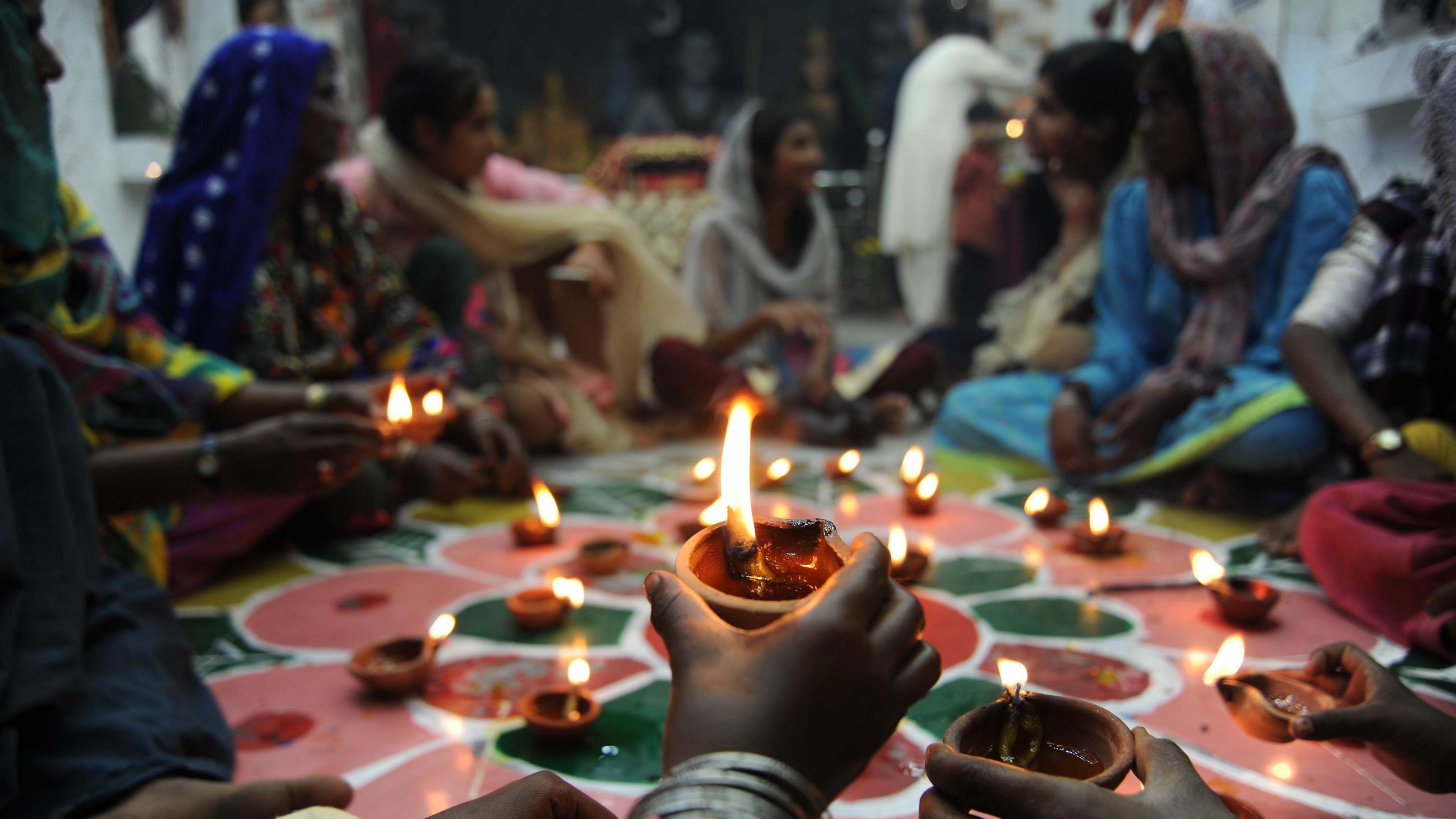 A small clay cup oil lamp with a flame is held among a circle of other similar lamps surrounded by women sitting cross-legged on a floor painted in a pink and green design