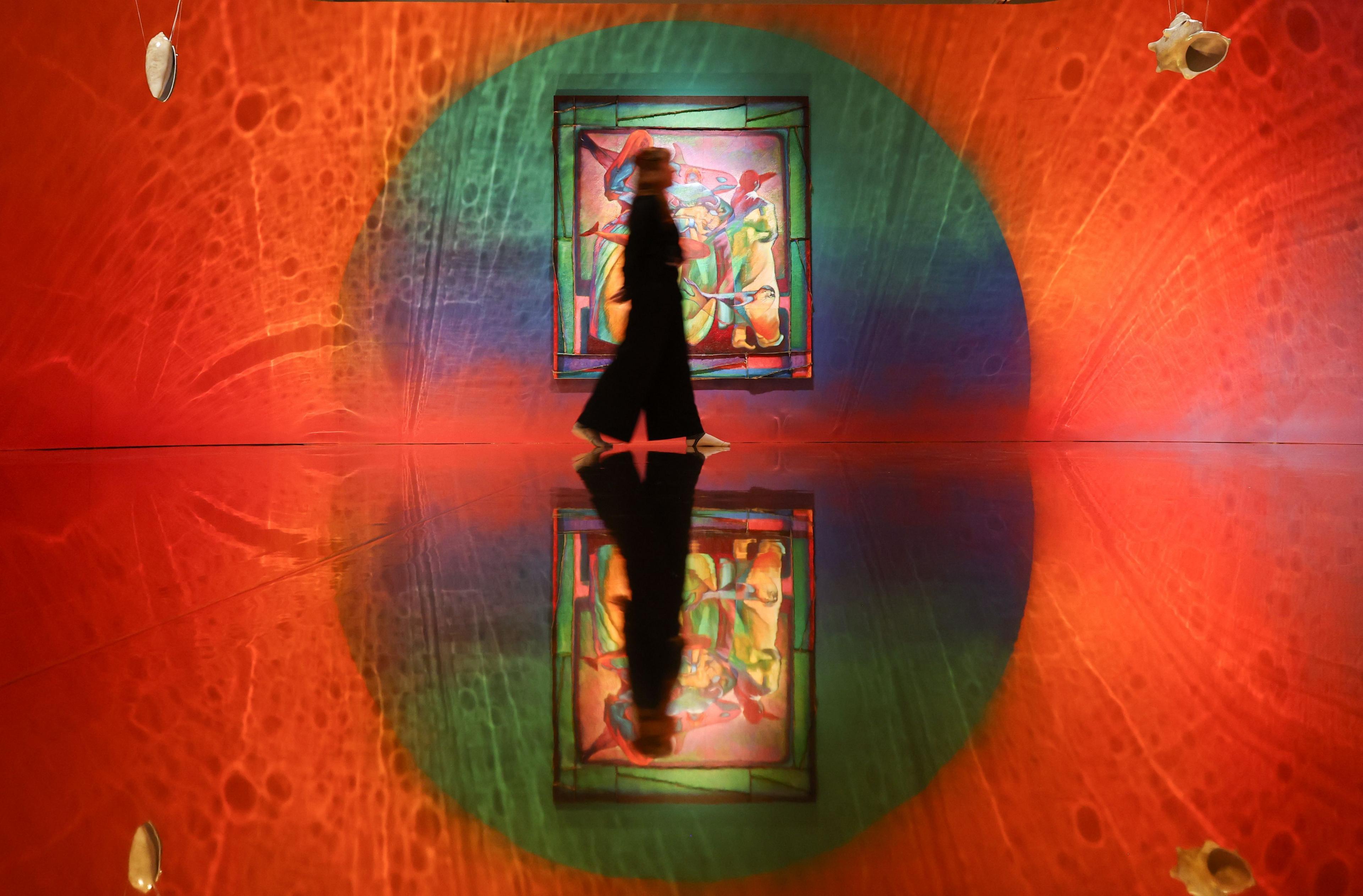 A woman walking past a large colourful painting in a gallery bathed mostly in red light and swirling shapes in Zadie Xa's installation