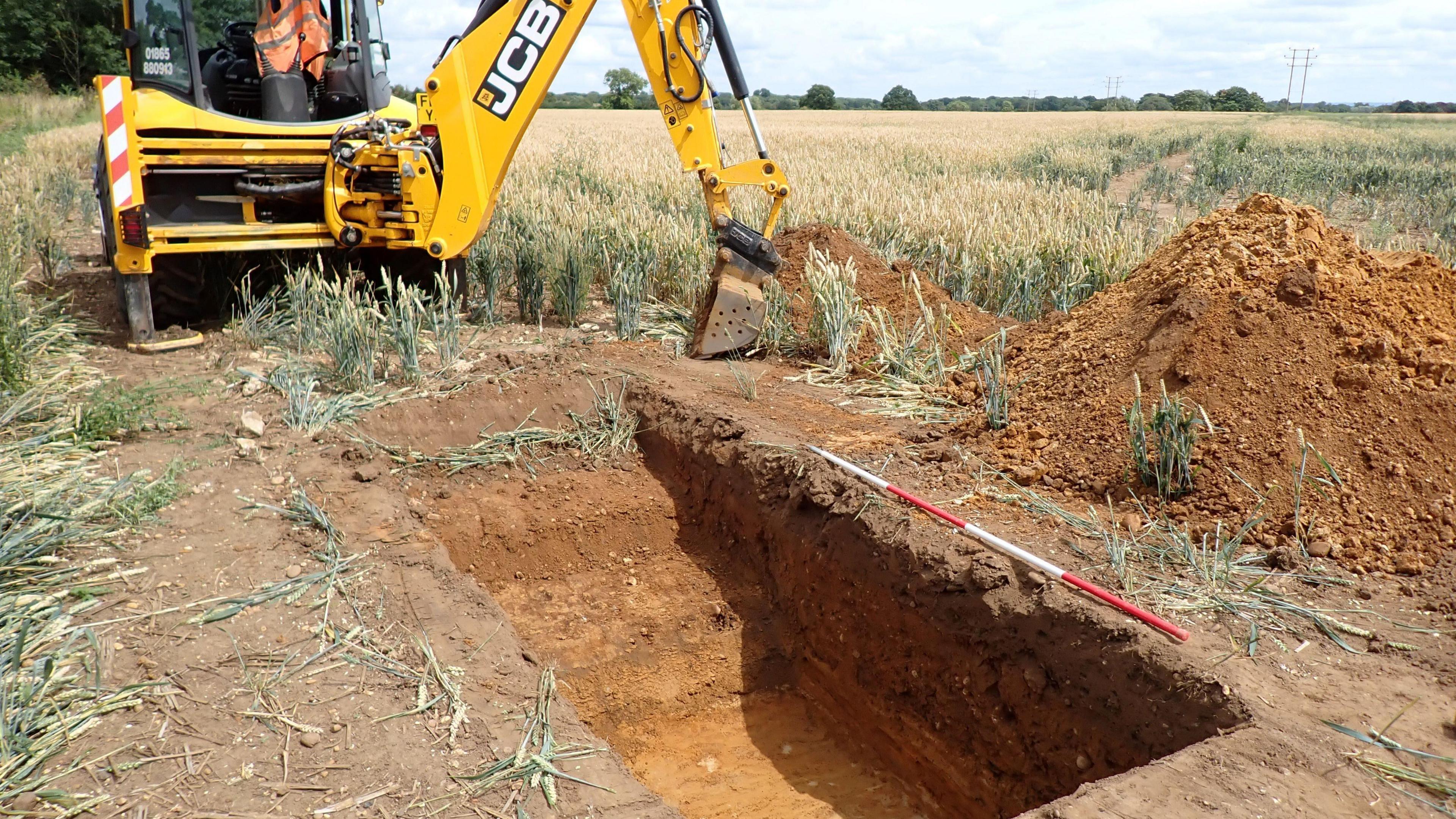 A yellow digger excavating a trench in a field. There is a red and white pole along the edge of the trench