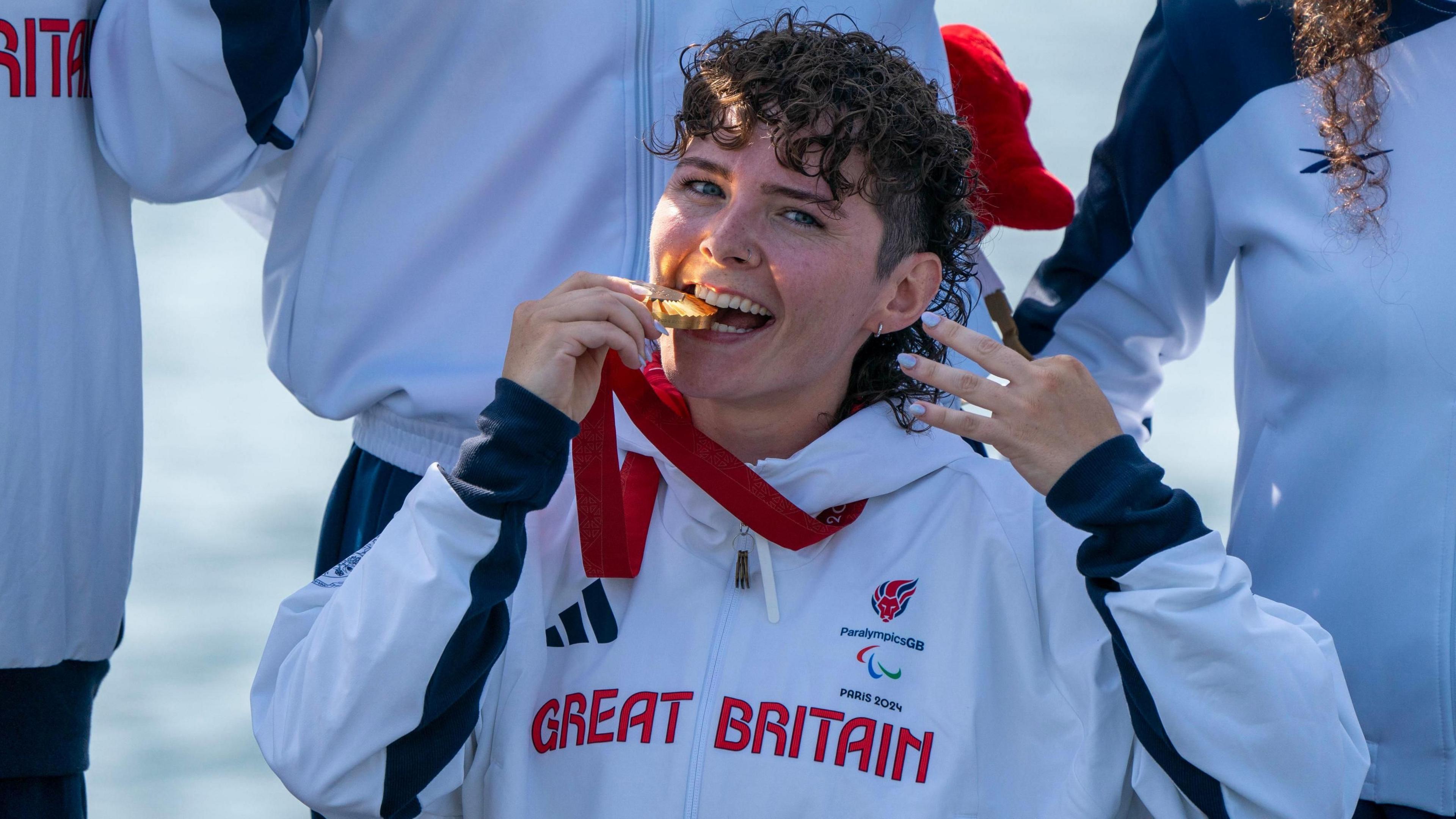 Lauren Rowles, a woman with short dark curly hair, bites down on a gold medal which is hanging around her neck. She is wearing a white hoodie emblazoned with "Great Britain", "Paralympics GB", and "Paris 2024". Others wearing similar outfits are standing around her, although their faces cannot be seen.