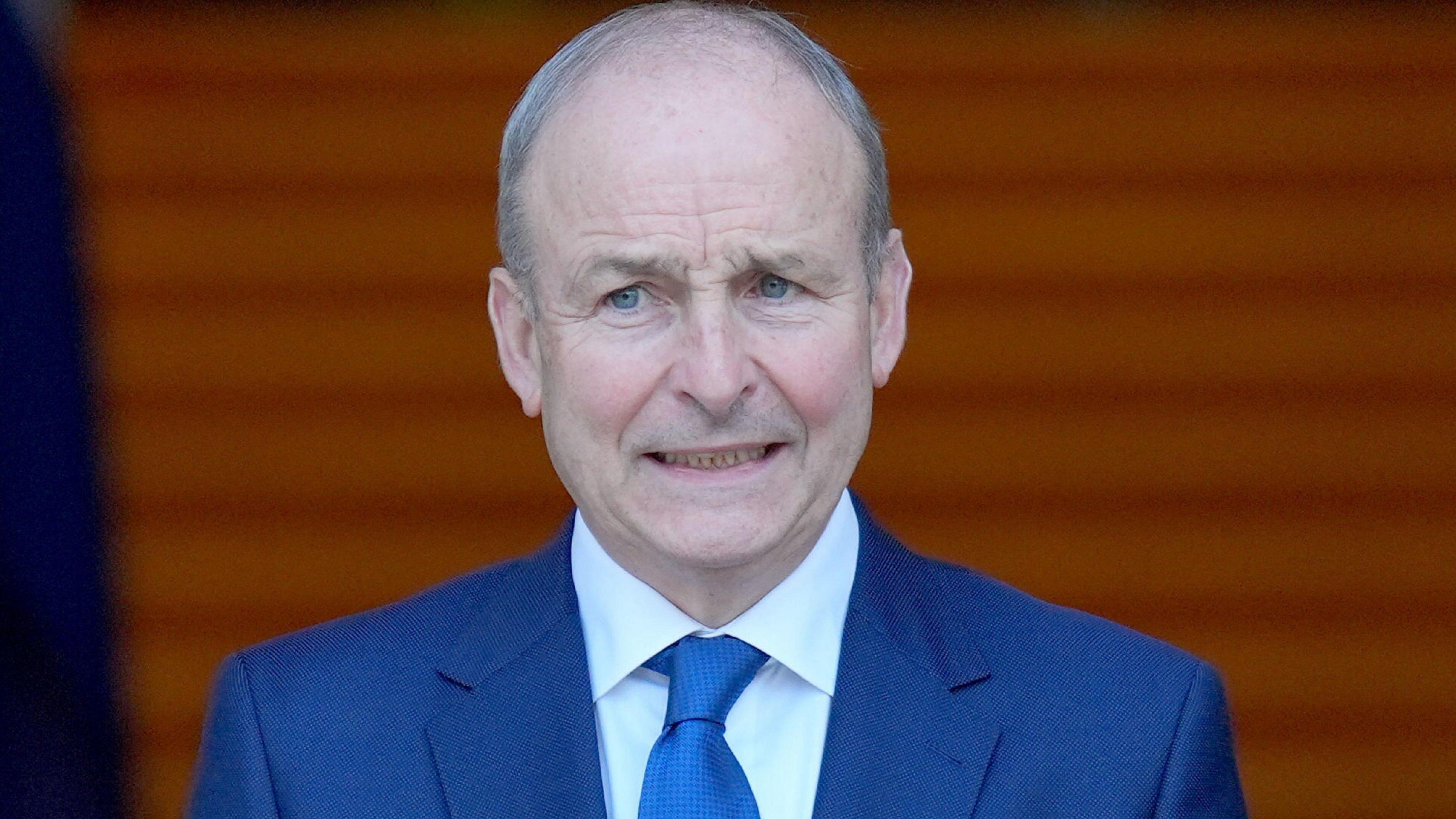 Micheál Martin has receding grey hair. He is wearing a blue suit and a blue shirt and tie.