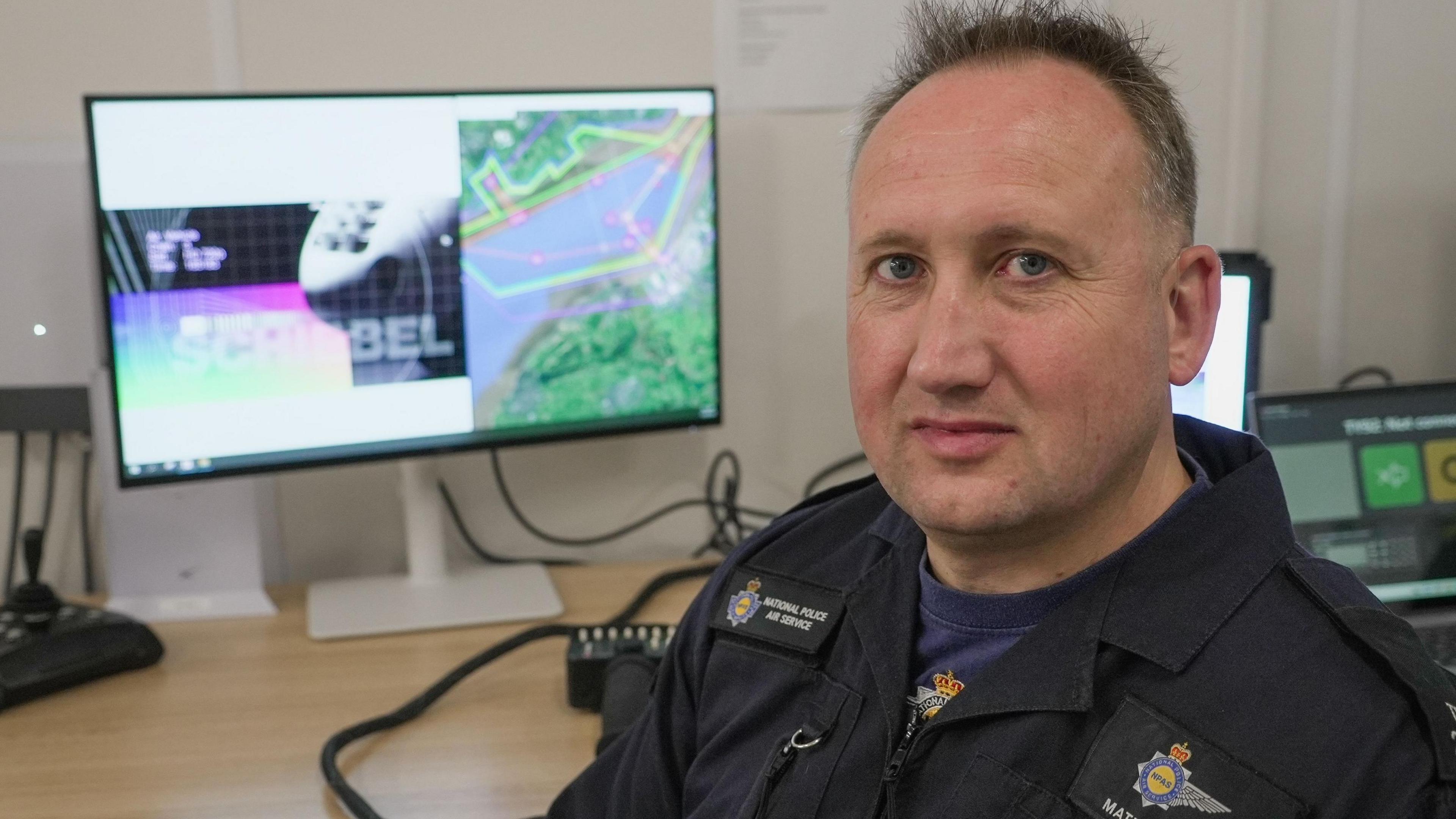A man with short hair looking into a camera. He is wearing a dark police NPAS uniform with a blue t-shirt underneath. He is sat at a desk with a computer monitor behind him, in an office setting.
