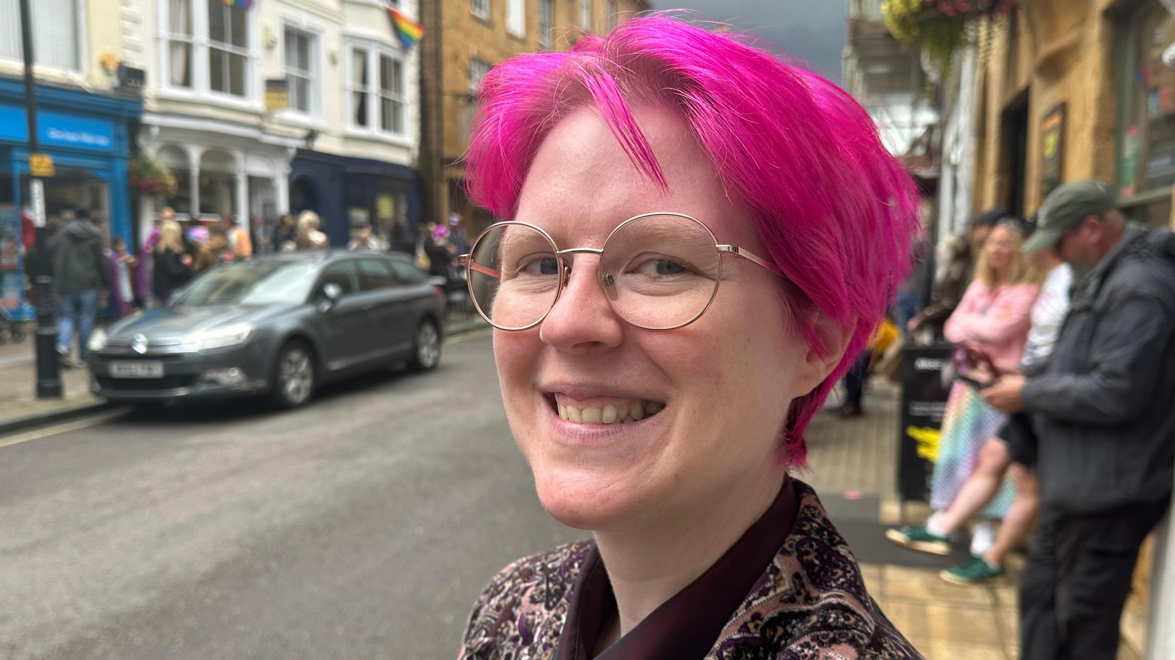 Lux Angelsong smiles for the camera on the street where the Pride parade is taking place. Lux has pink hair and circular glasses.