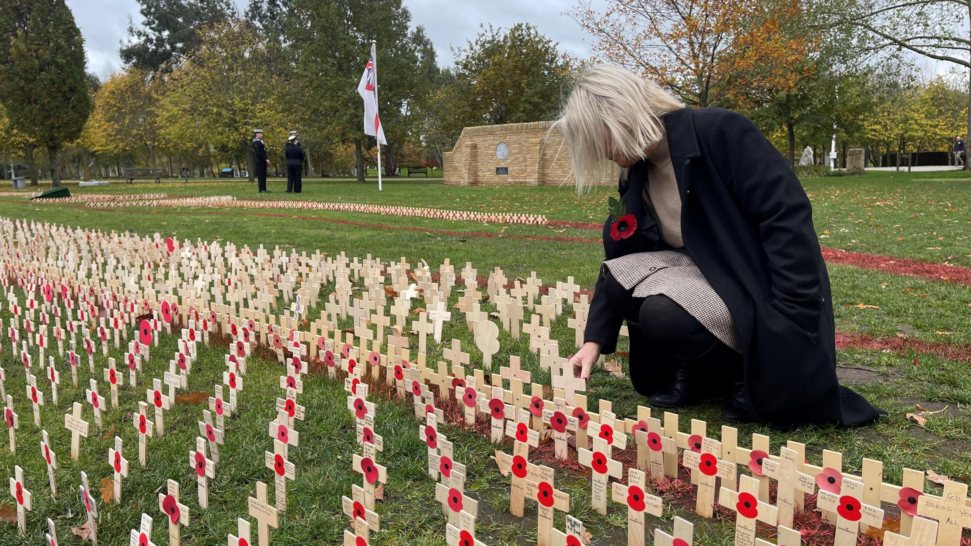 A woman is kneeling down to read one of the hundreds of wooden crosses on display. They each bear the image of a red poppy and a handwritten note and are placed in the grass. There are military memorials and people in service uniform in the background.