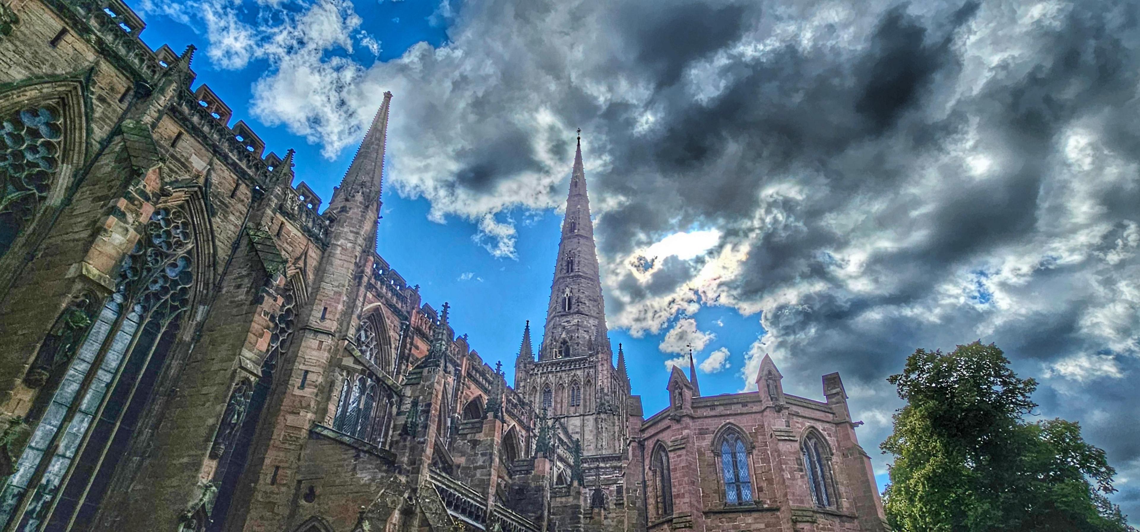 The spire and roof of Lichfield Cathedral, set against black cloud massing across the sun, with blue sky still visible to one side.