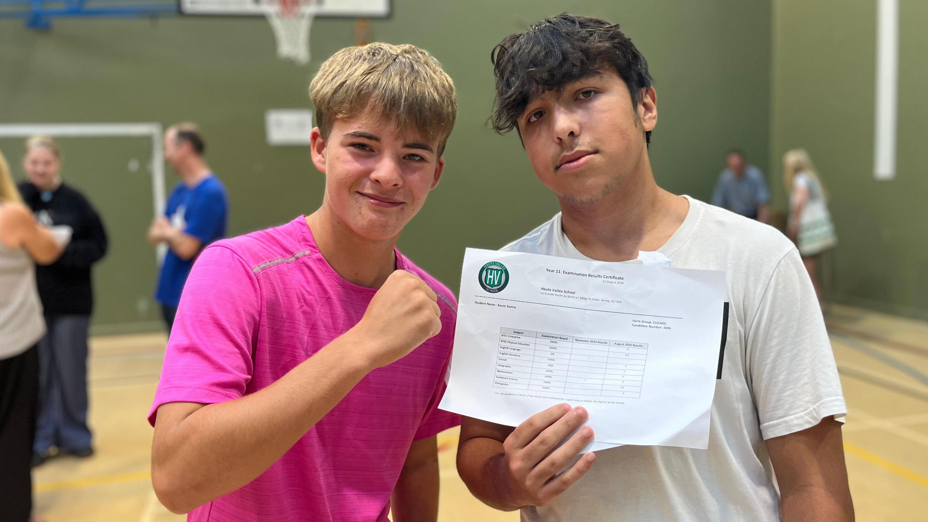 Jack Peron is raising his fist and smiling and Kevin Aveiar is holding his GCSE results paperwork up to the camera. They are in a school sports hall.