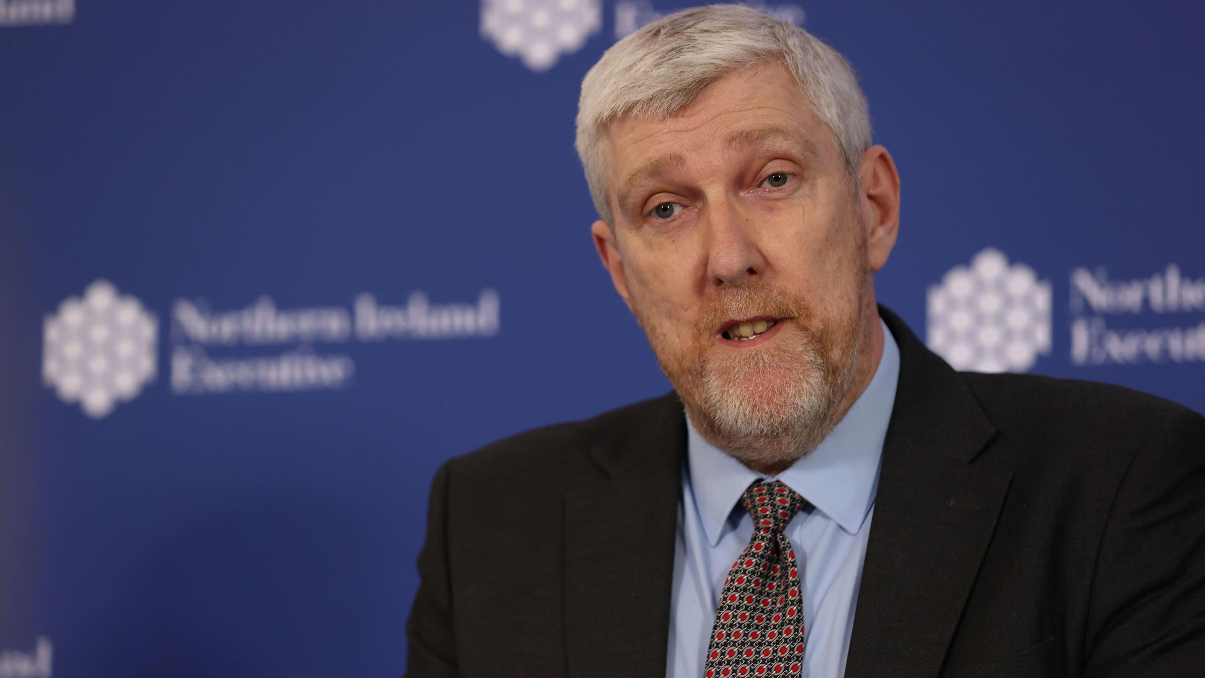 John O'Dowd standing in front of a blue wall that sasy Northern Ireland Executive, he has white hair and is wearing a black suit with light blue shirt and red polka dot tie