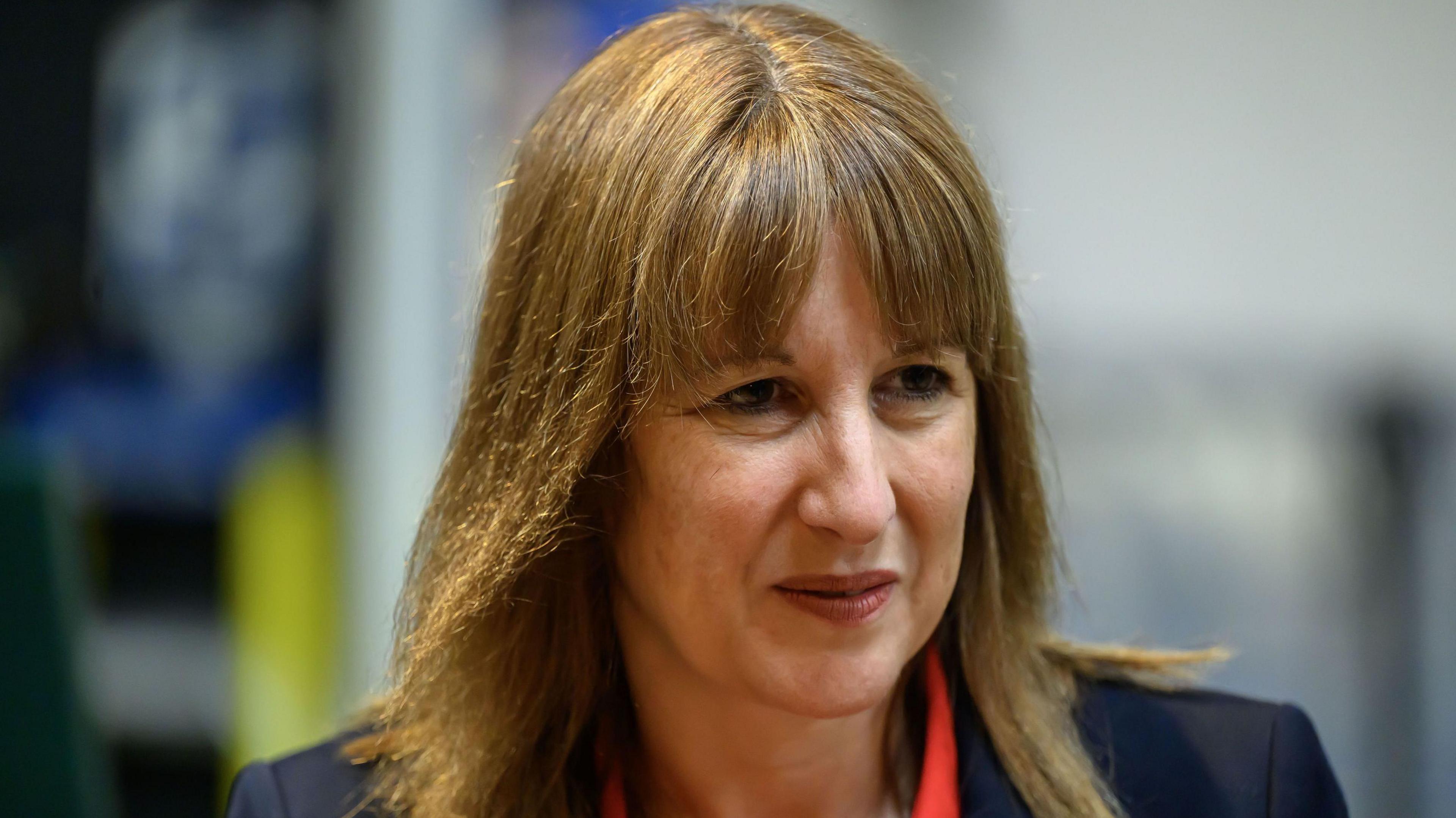 Chancellor Rachel Reeves wearing a dark blazer over a bright red blouse, in front of a blurred background suggesting an office or meeting setting