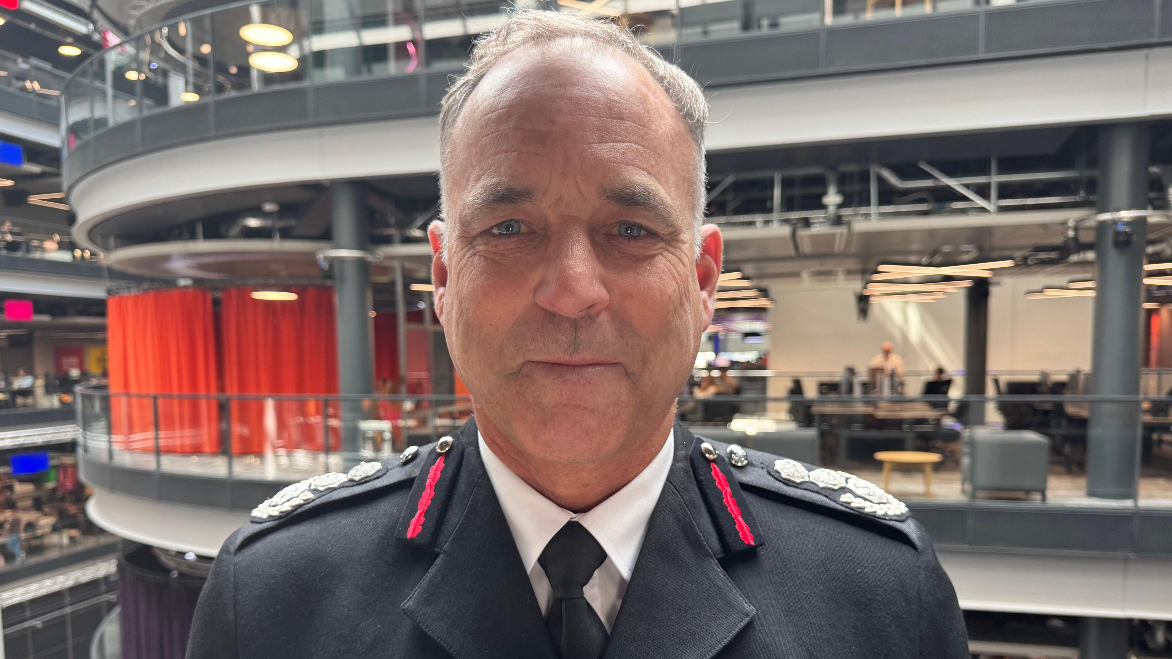 Fin Monahan pictured stood on a balcony inside BBC Wales' headquarters in Cardiff. He wears his full fire uniform, complete with a black suit, white shirt and tie. He has blue eyes, short silver hair and smiles at the camera.