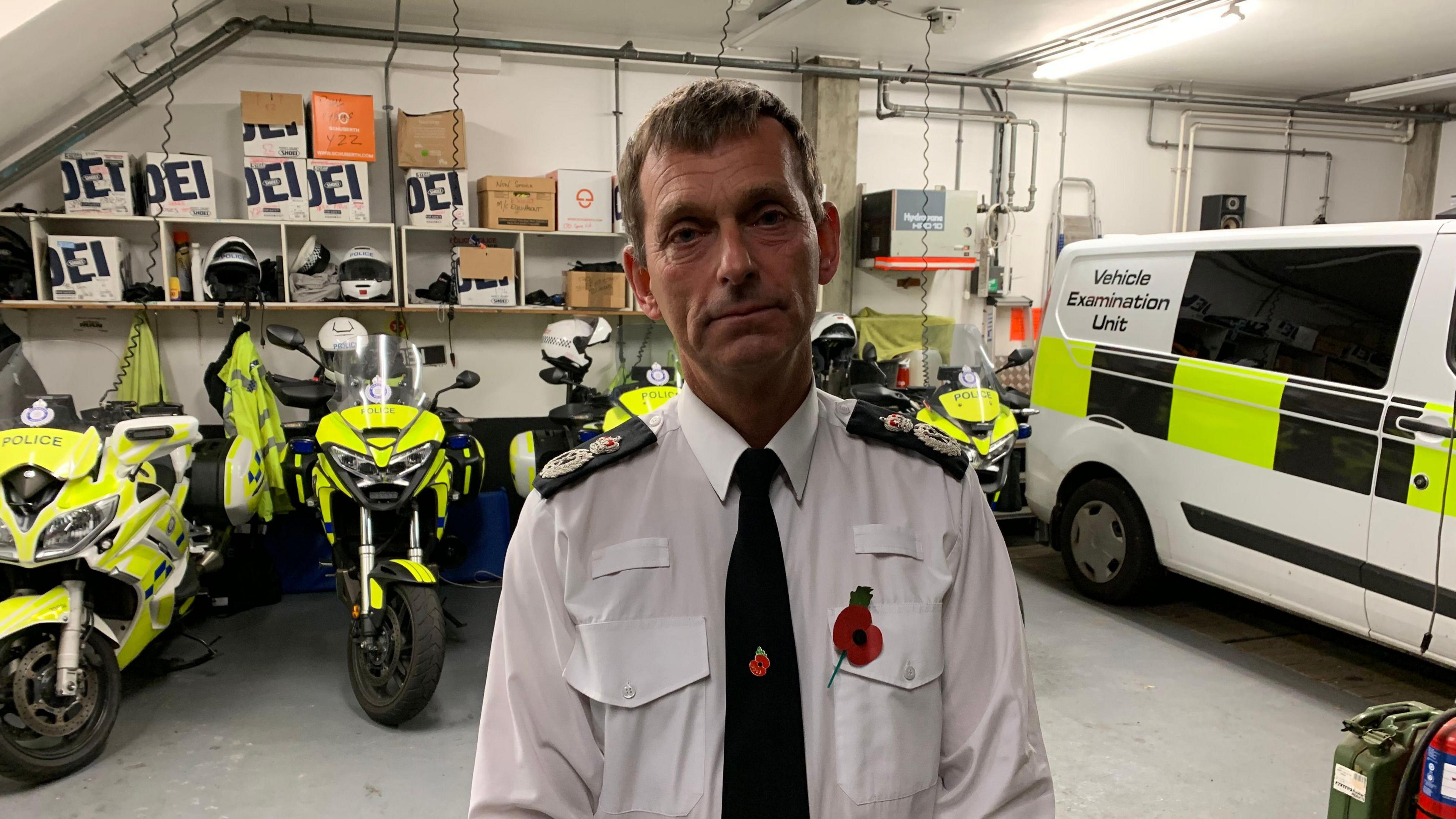 Close up of former police chief Ruari Hardy. Mr Hardy is wearing a white police uniform shirt. He is wearing a black tie and two red poppy pins. He is standing in front of a police vehicle and multiple police motorbikes.