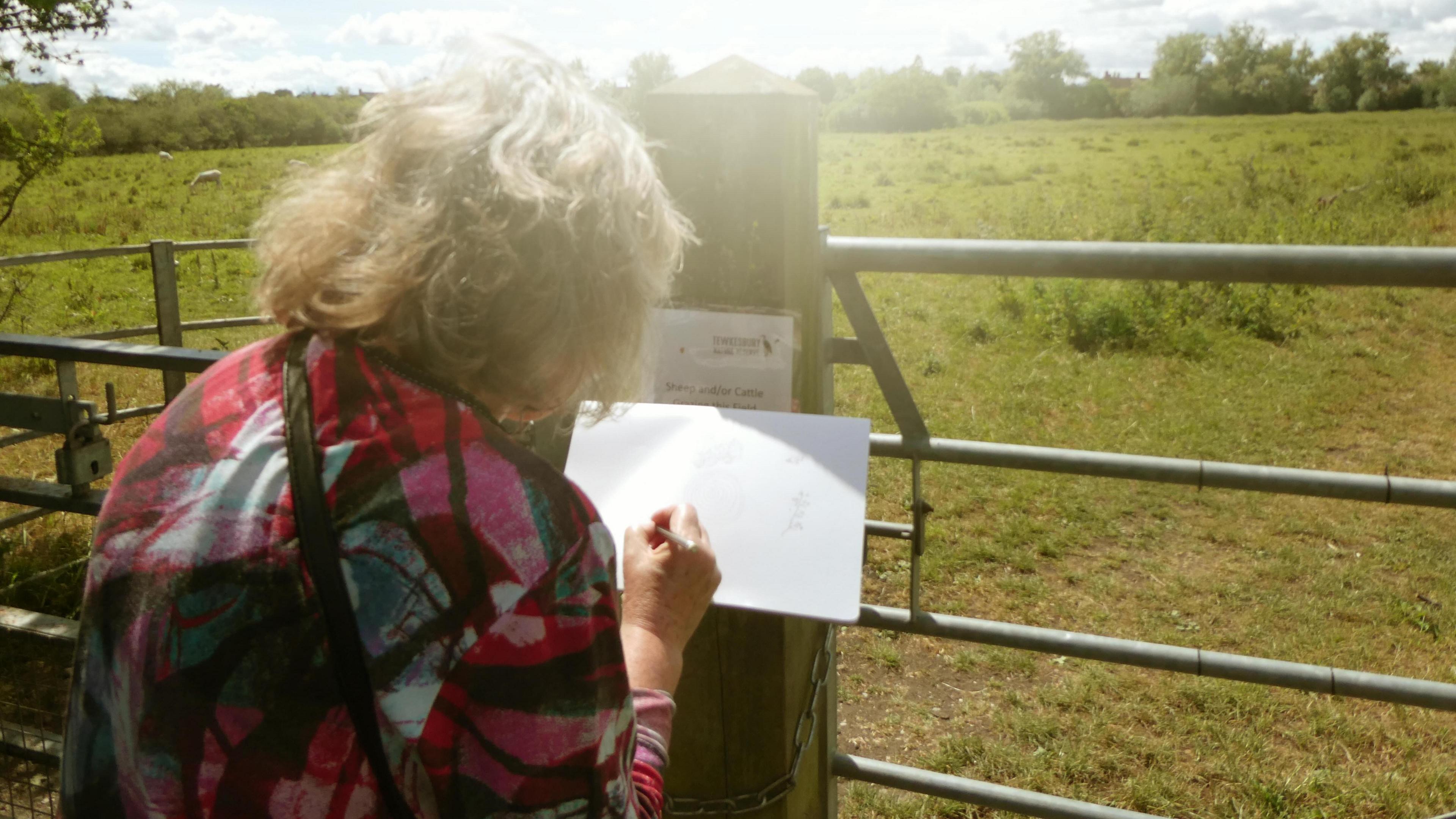An older woman holds the Tewkesbury Wellbeing Walks map in front of a gate, with greenery in the background.