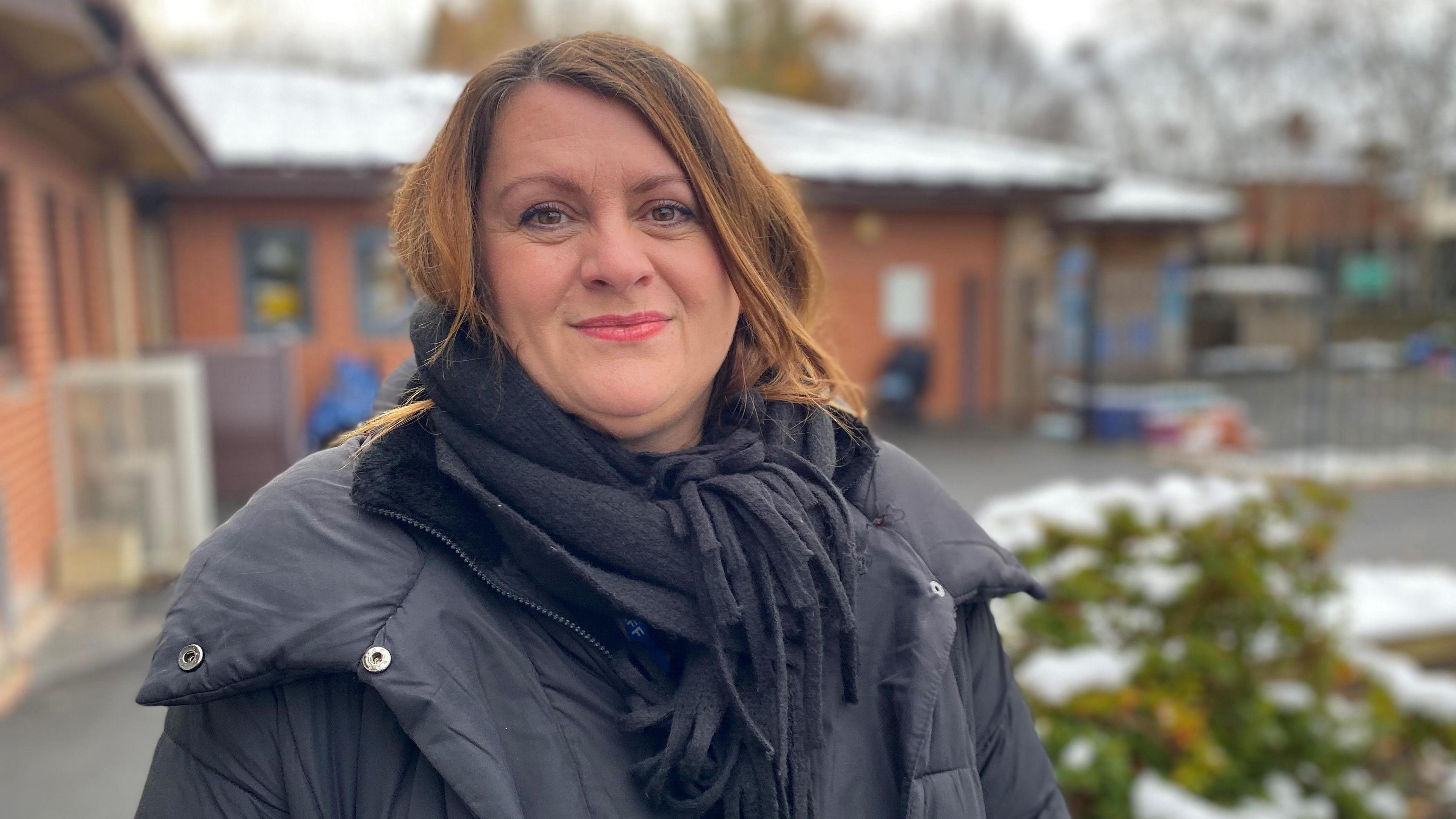 Ms Watson wears a black scarf and coat as she stands in the snowy school grounds. She wears red lipstick and has dyed copper hair.