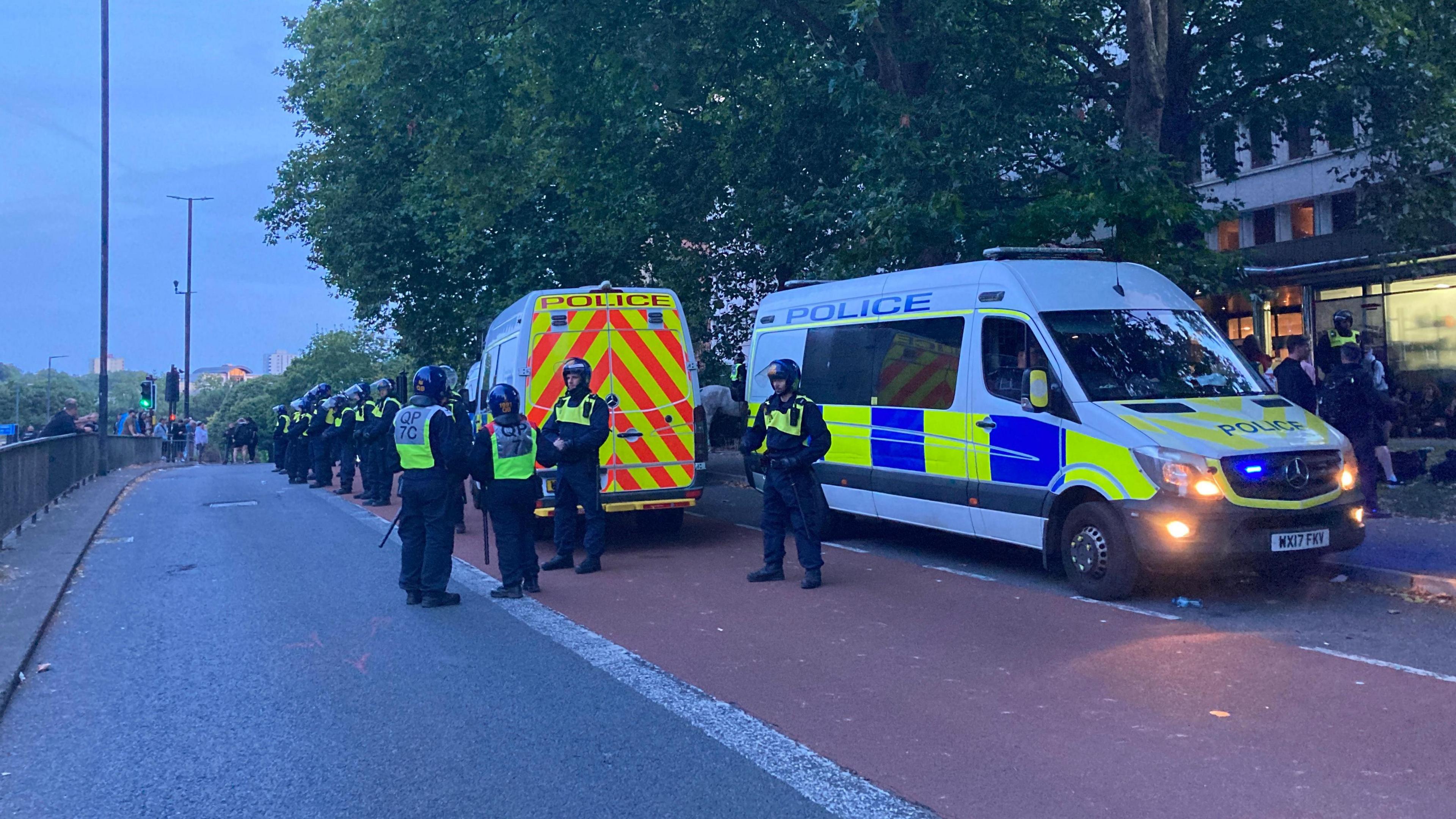 Police vans parked in a bus lane with a long line of police officers wearing helmets and high visibility jackets