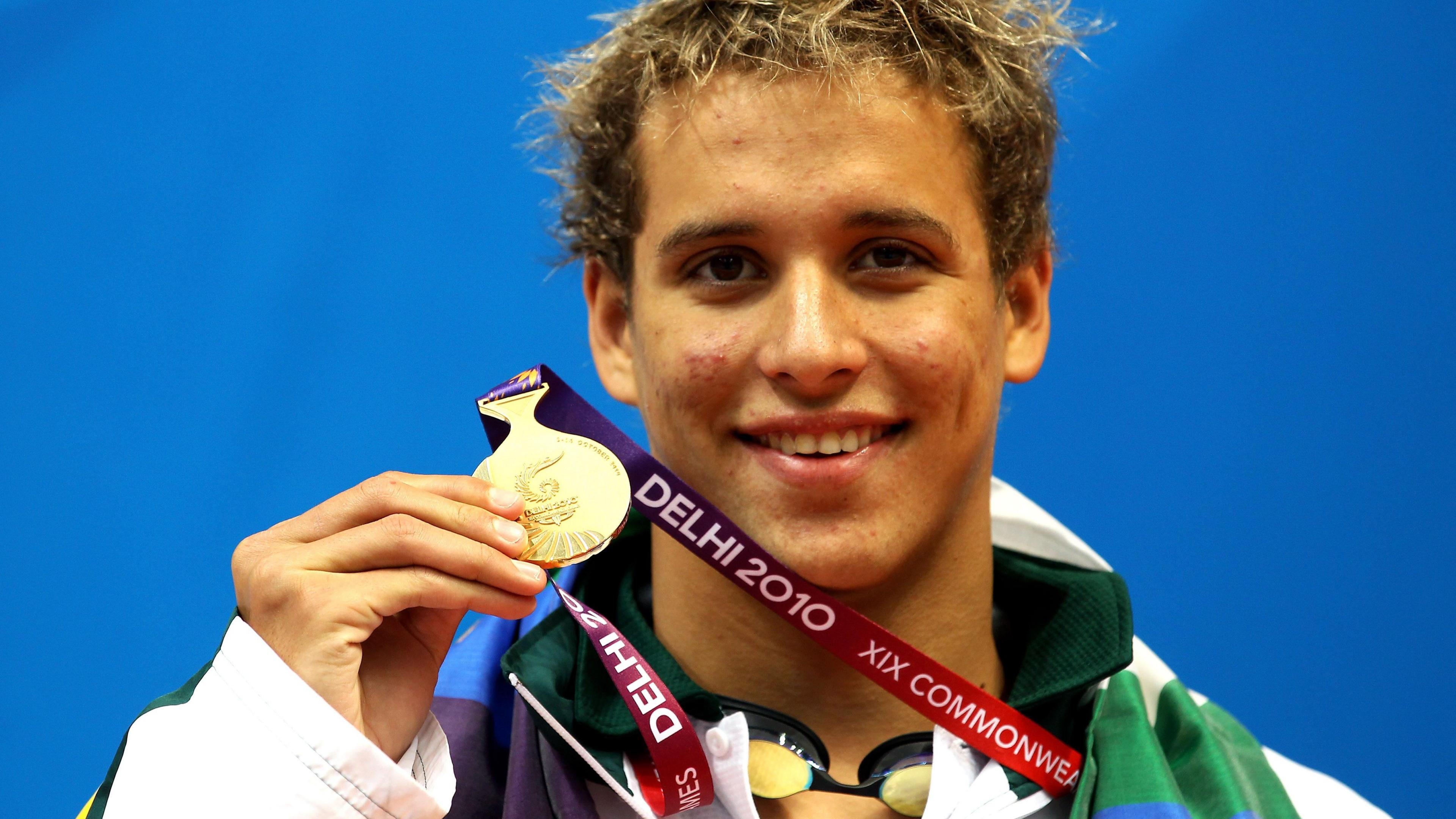 A head and shoulders shot of Chad le Clos shows him smiling as he holds up a gold medal on a dark red Delhi 2010 ribbon with his right hand. He wears a predominantly white and green tracksuit jacket and a pair of swimming goggles are visible around his neck