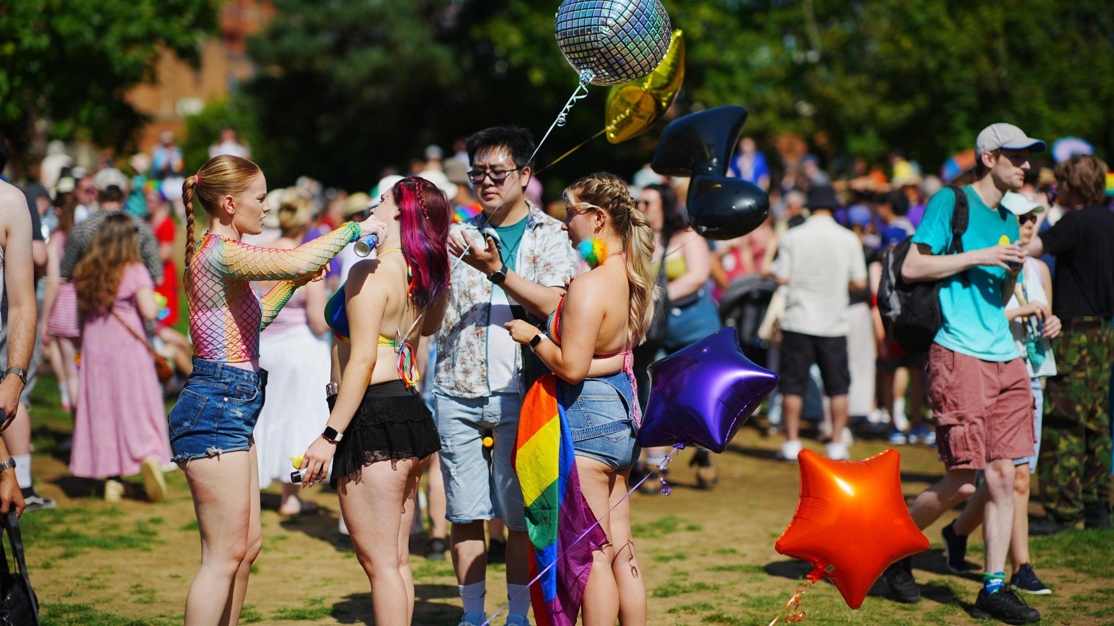 A crowd of young people in colourful clothing gather in a field in front of a large gathering of people with pride flags and balloons.