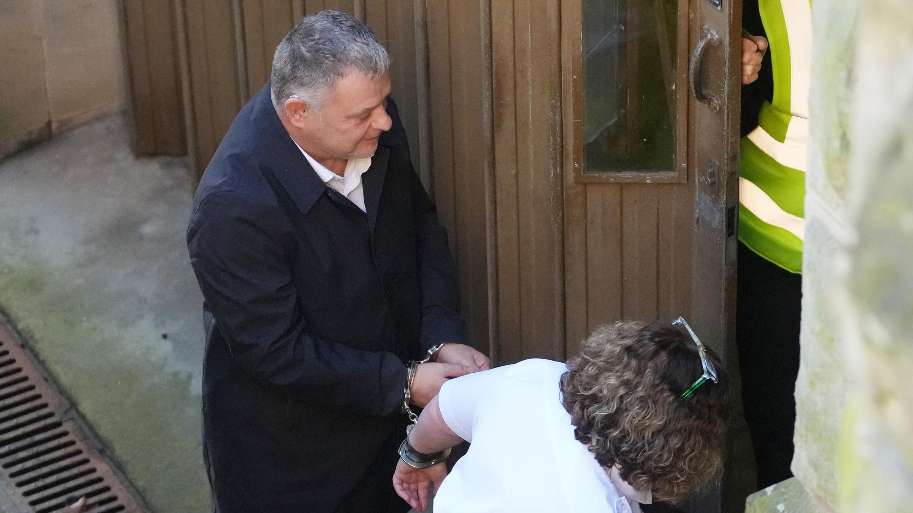 Mike Amesbury, wearing a black coat and white shirt, enters the court building handcuffed to a partially obscured prison officer.