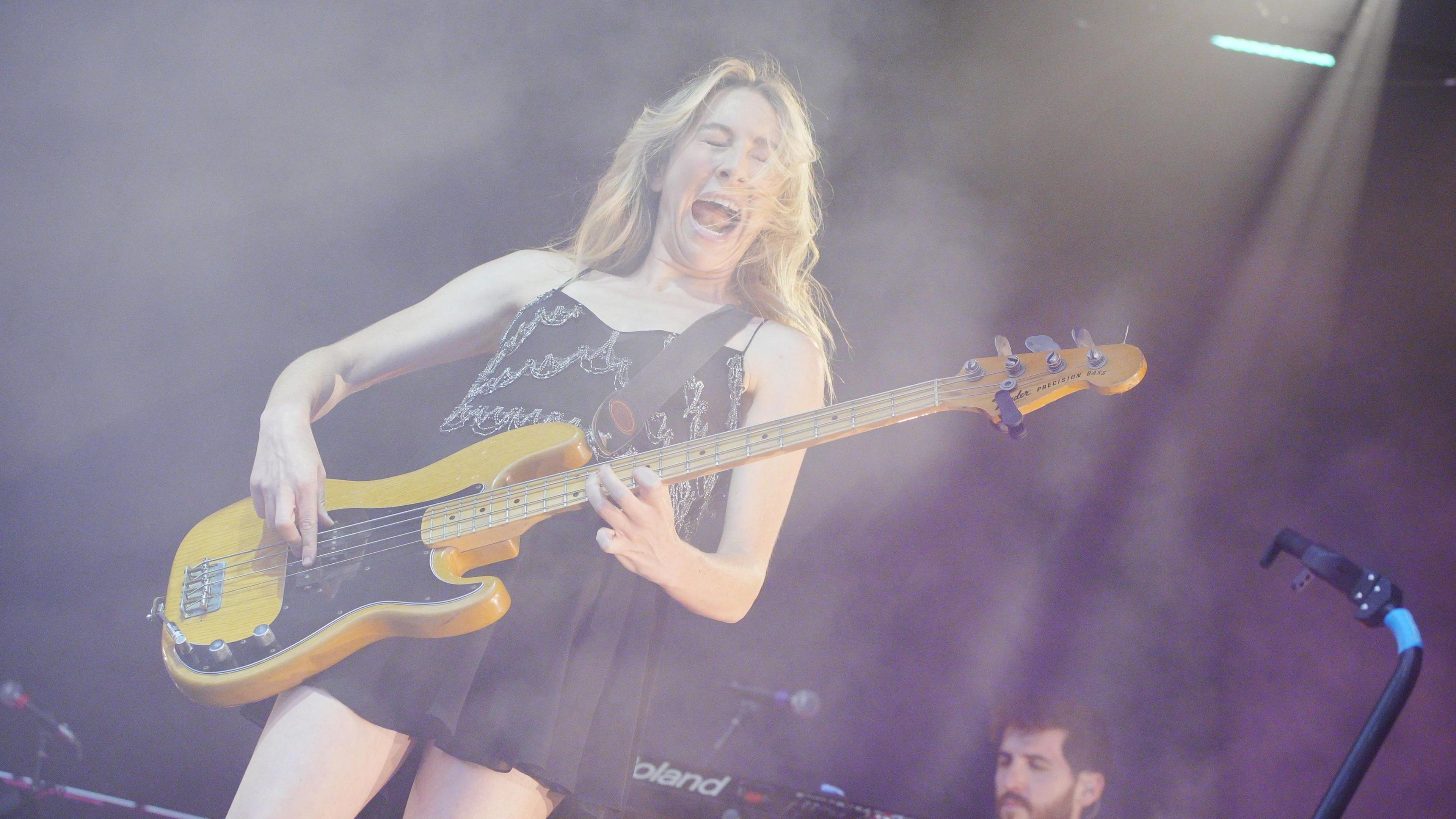 A woman performs on stage wearing a black dress. She is playing an electric guitar, which is yellow and black. Her long blonde hair is partially in her face as she moves. She has her eyes closed and appears to be singing or shouting, with her mouth wide open.
