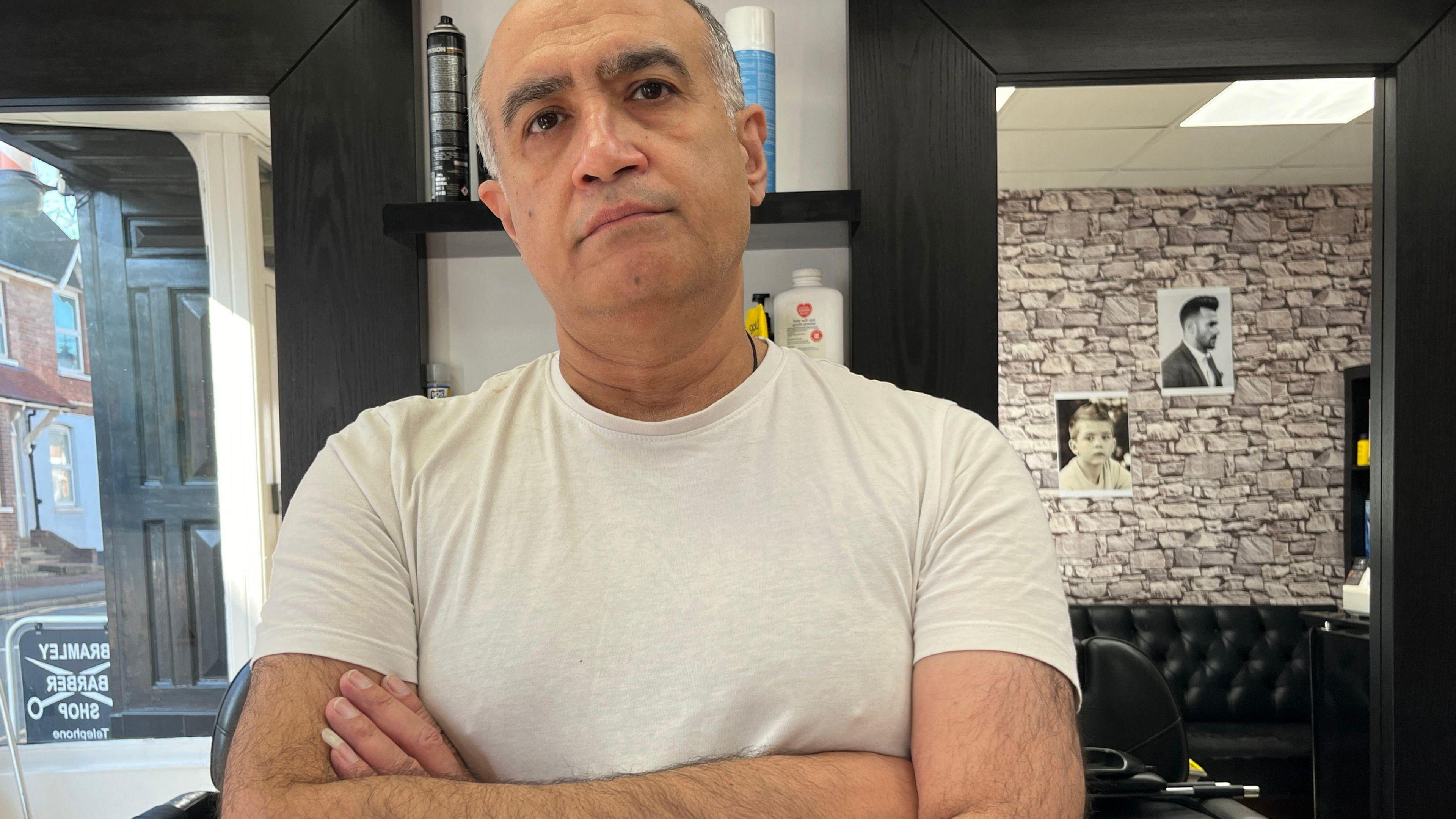 Mehran Shafaie is standing in a white t-shirt with his arms folded in his barber shop