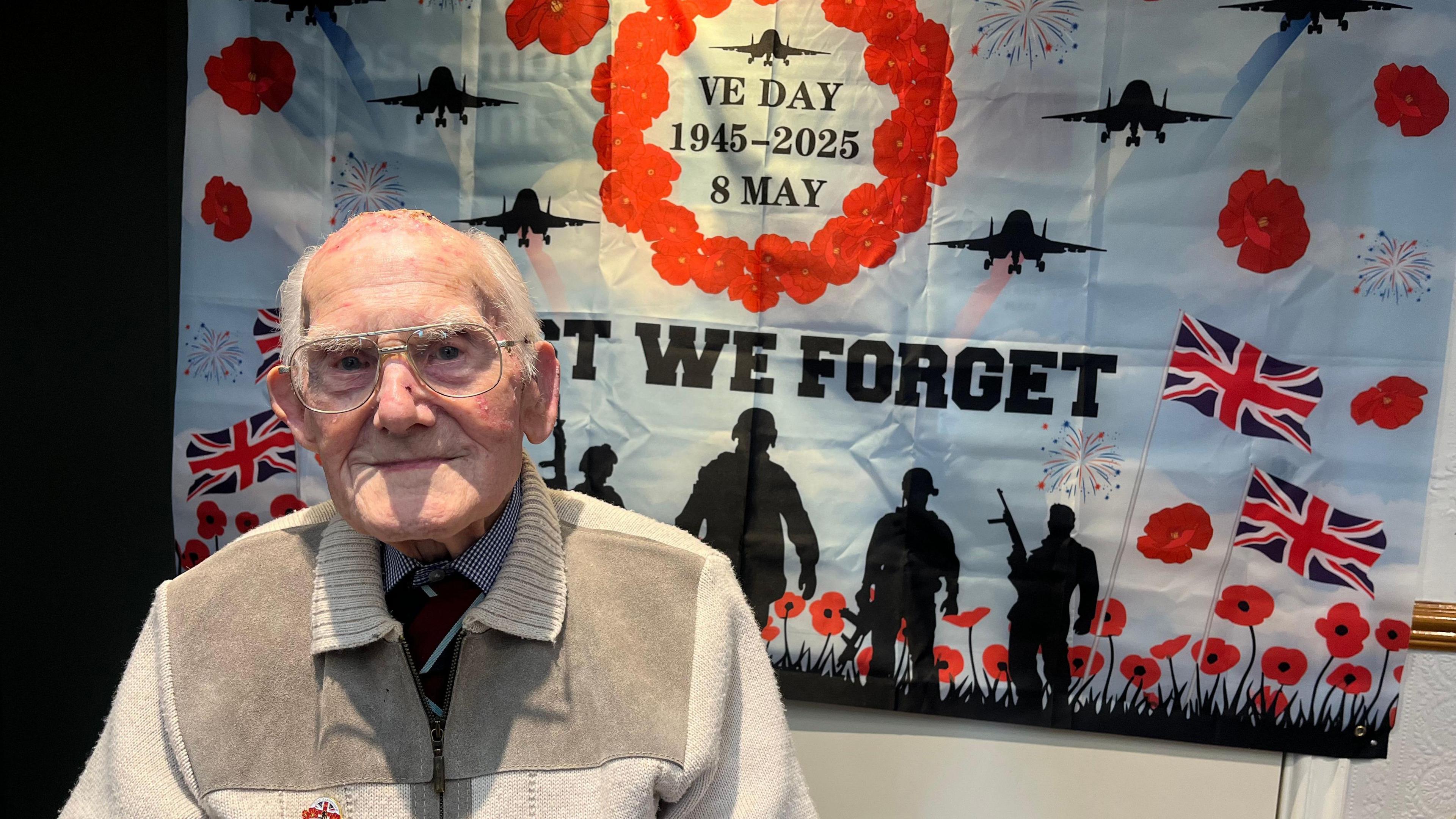 An old man with glasses, looks at the camera, as he sits in front of large poster. The poster has a blue sky background with red poppies on it and silhoutees of soldiers, with British flags. It reads 'lest we forget' and 'VE DAY 1945-2025- 8 MAY'