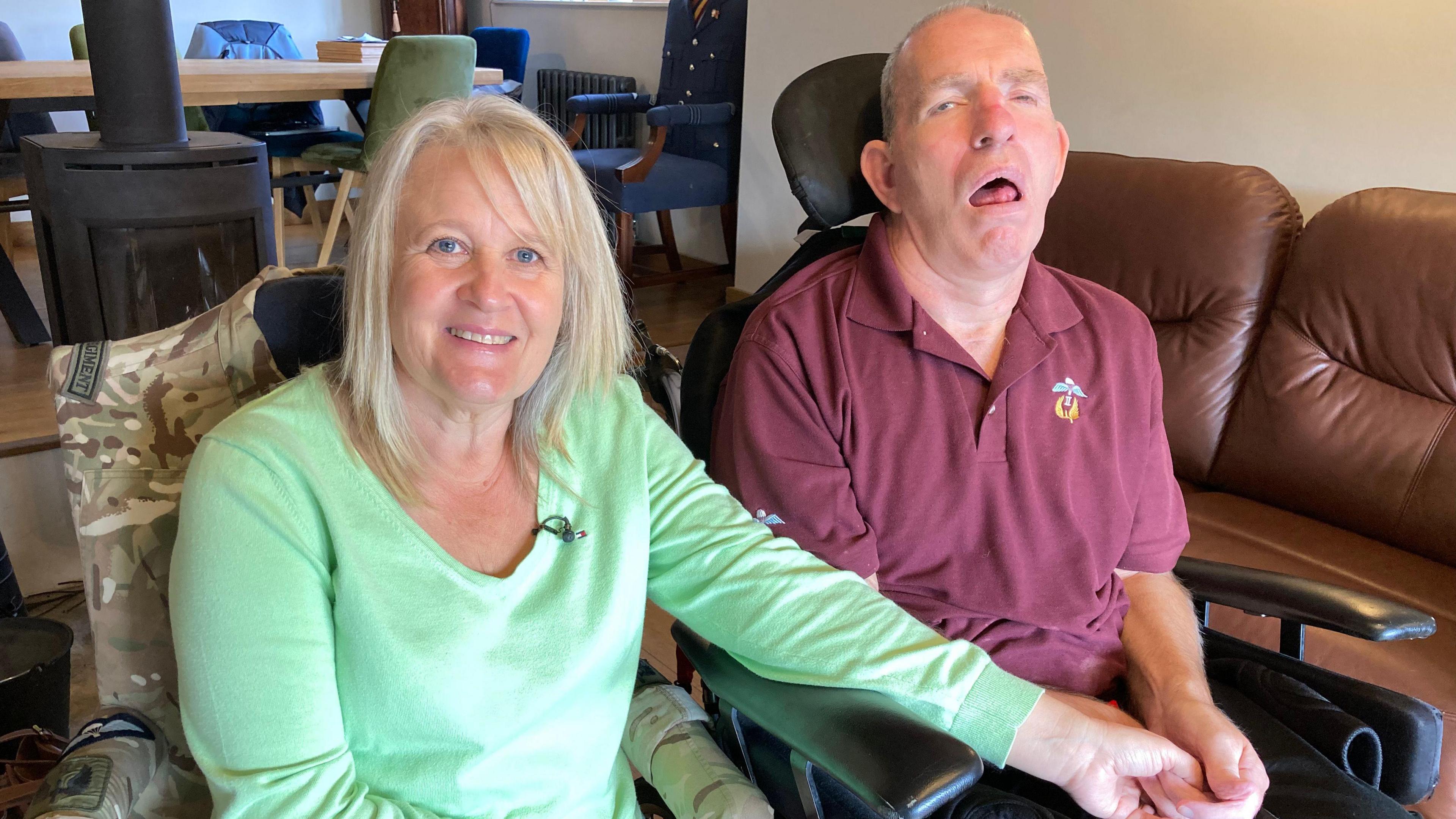 Sharon with Andy in their home. Sharon is smiling looking at the camera in a mint green top holding Andy's hand whilst he is sat in a wheelchair