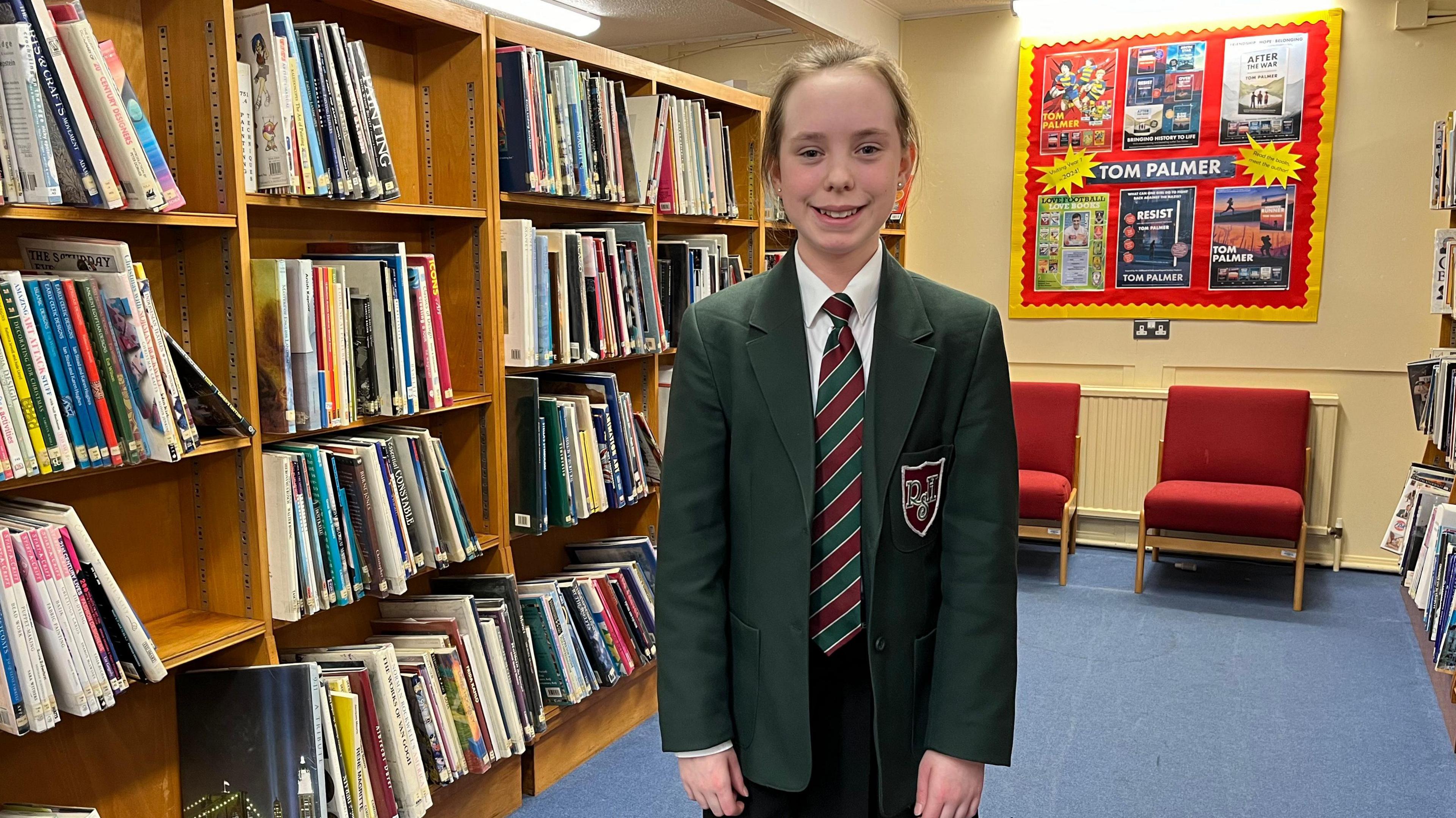 Eve stands in the school library surrounded by books. She is wearing a dark green blazer and striped school tie. 