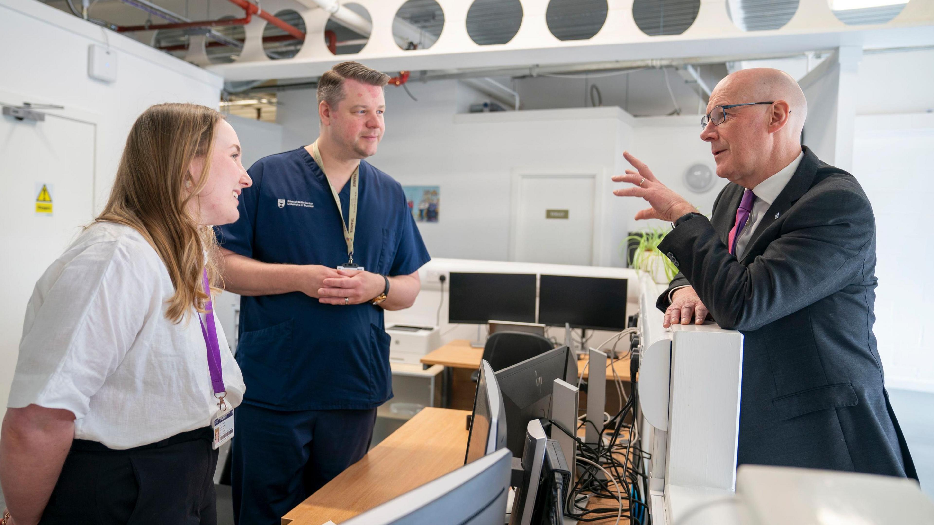 A woman and a man in medical scrubs speak to John Swinney in a suit leaning over a desk of computers