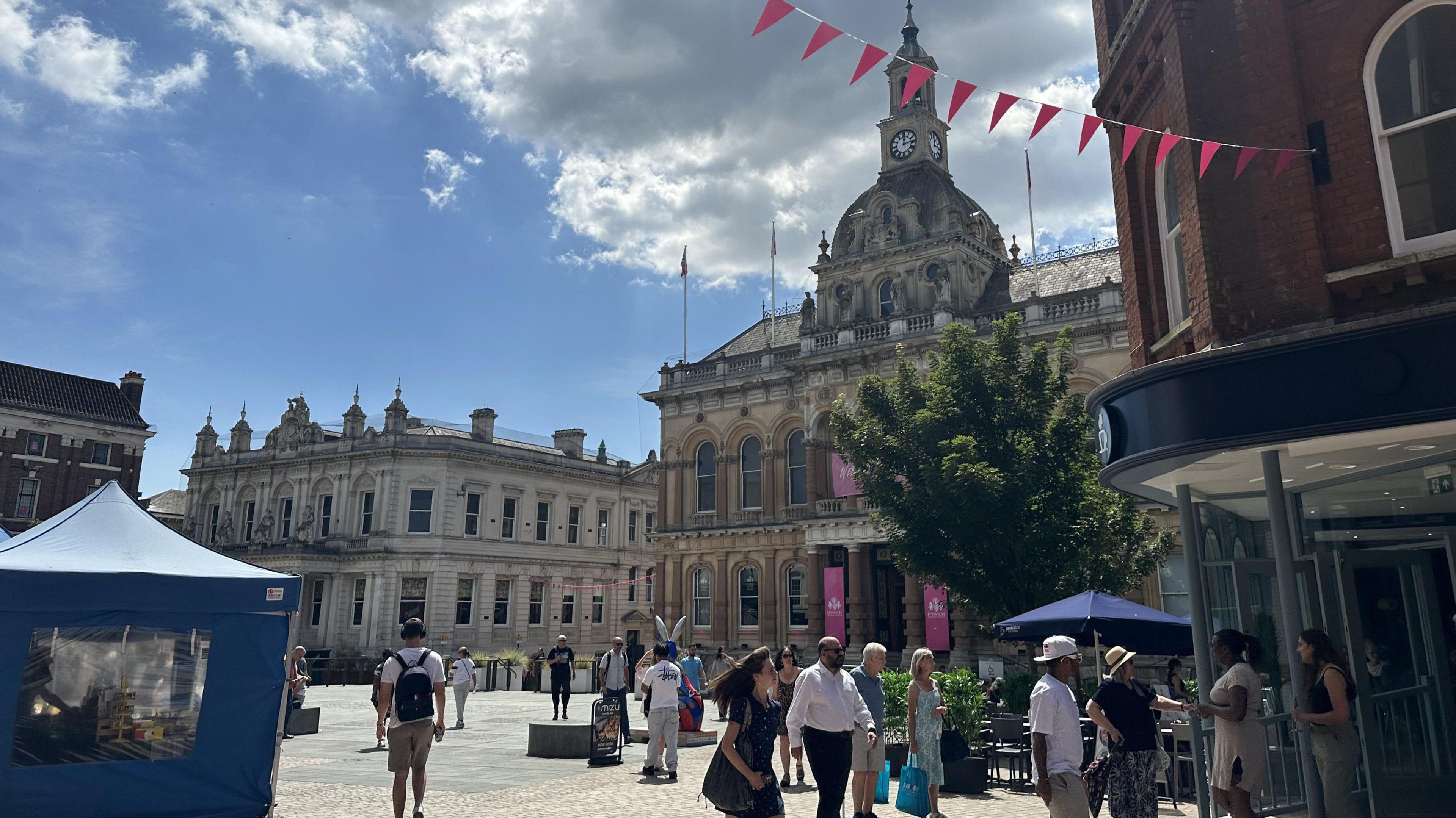 A general view of Ipswich town centre on a sunny day. A large town hall sits toward the centre right of the image with a large square outside of it that people walk about it. Pink bunting can be seen tied up between surrounding buildings.