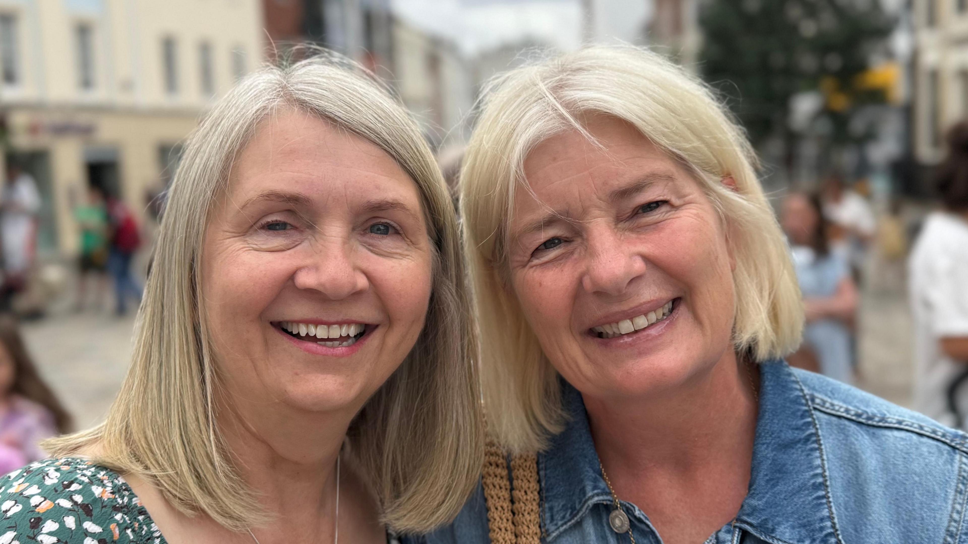 Two women standing side-by-side and smiling straight at the camera. They are standing in a city centre. The woman on the left is wearing a green patterned blouse and the other a blue denim jacket.
