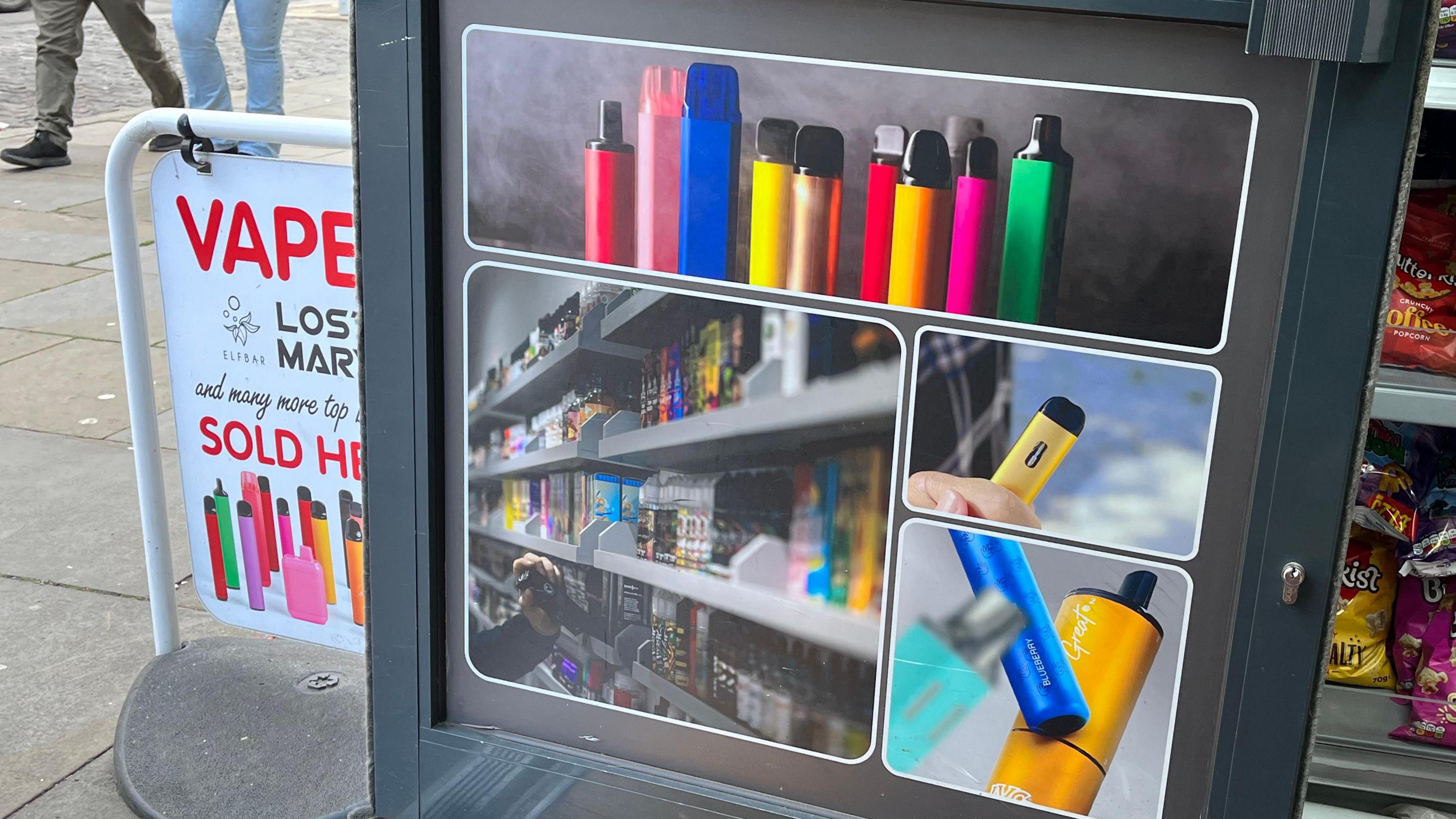 The lower half of a shop door with pictures of brightly-coloured vapes on it. Just outside the door is a sign that reads in red letting 'Vape - Lost Mary and many more top brands sold here' on a white background.