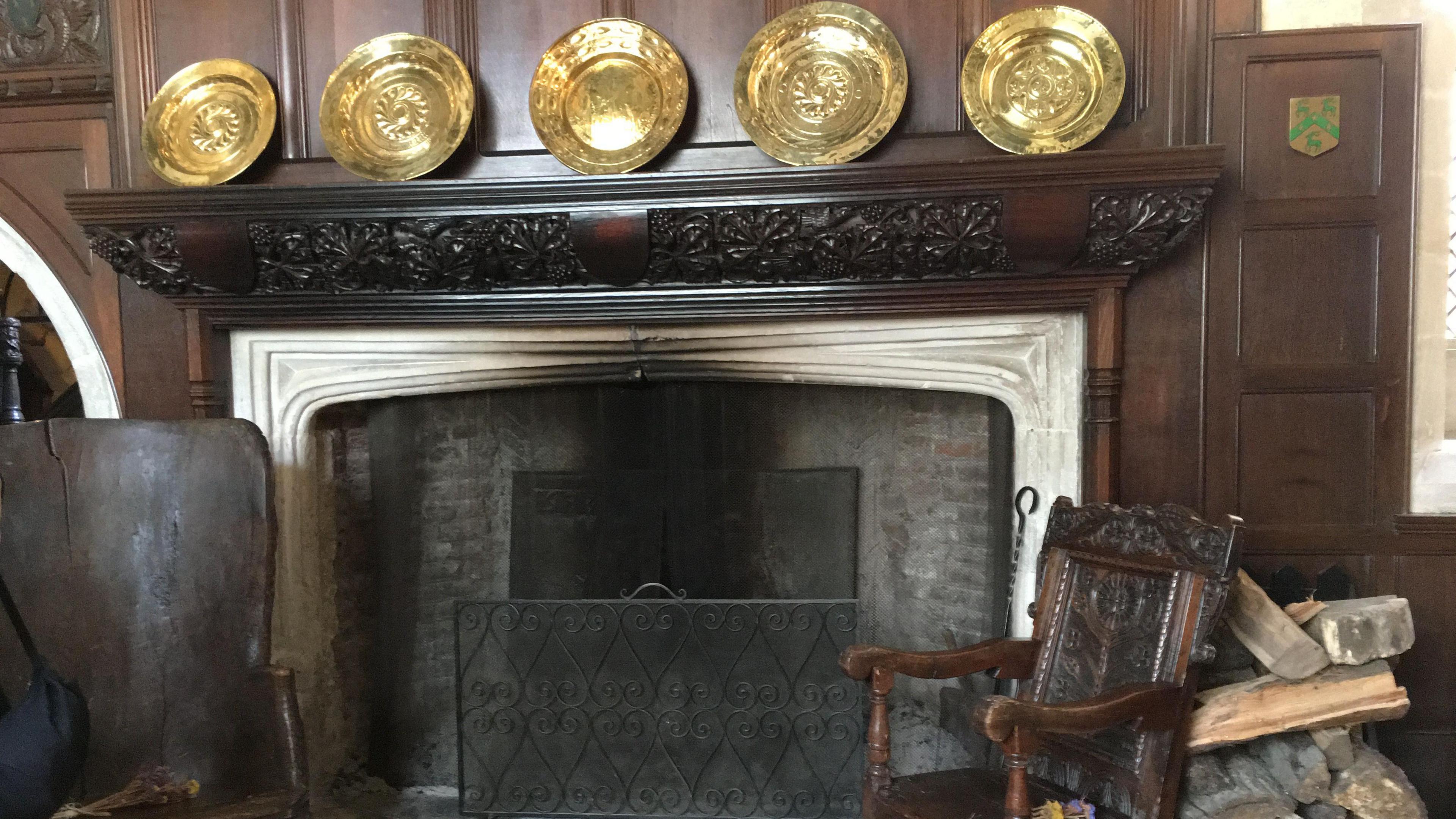 A large stone fireplace with dark carved wood surround and panelling, mantelpiece has gold-coloured decorative platters and there are two dark wood chairs in front.