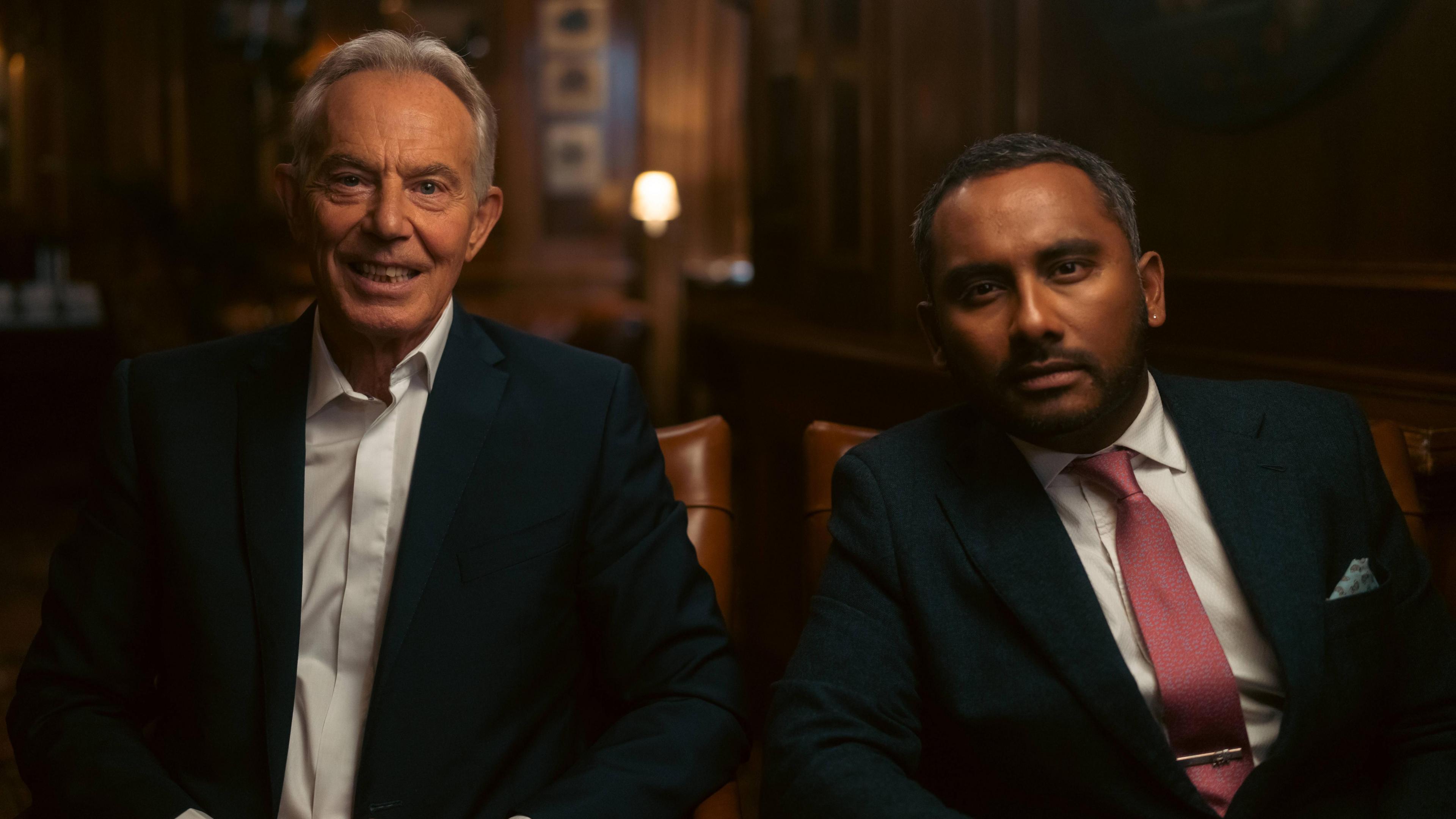 Tony Blair and Amol Rajan are sat in a dimly lit room. They are both wearing suits and looking directly at the camera.