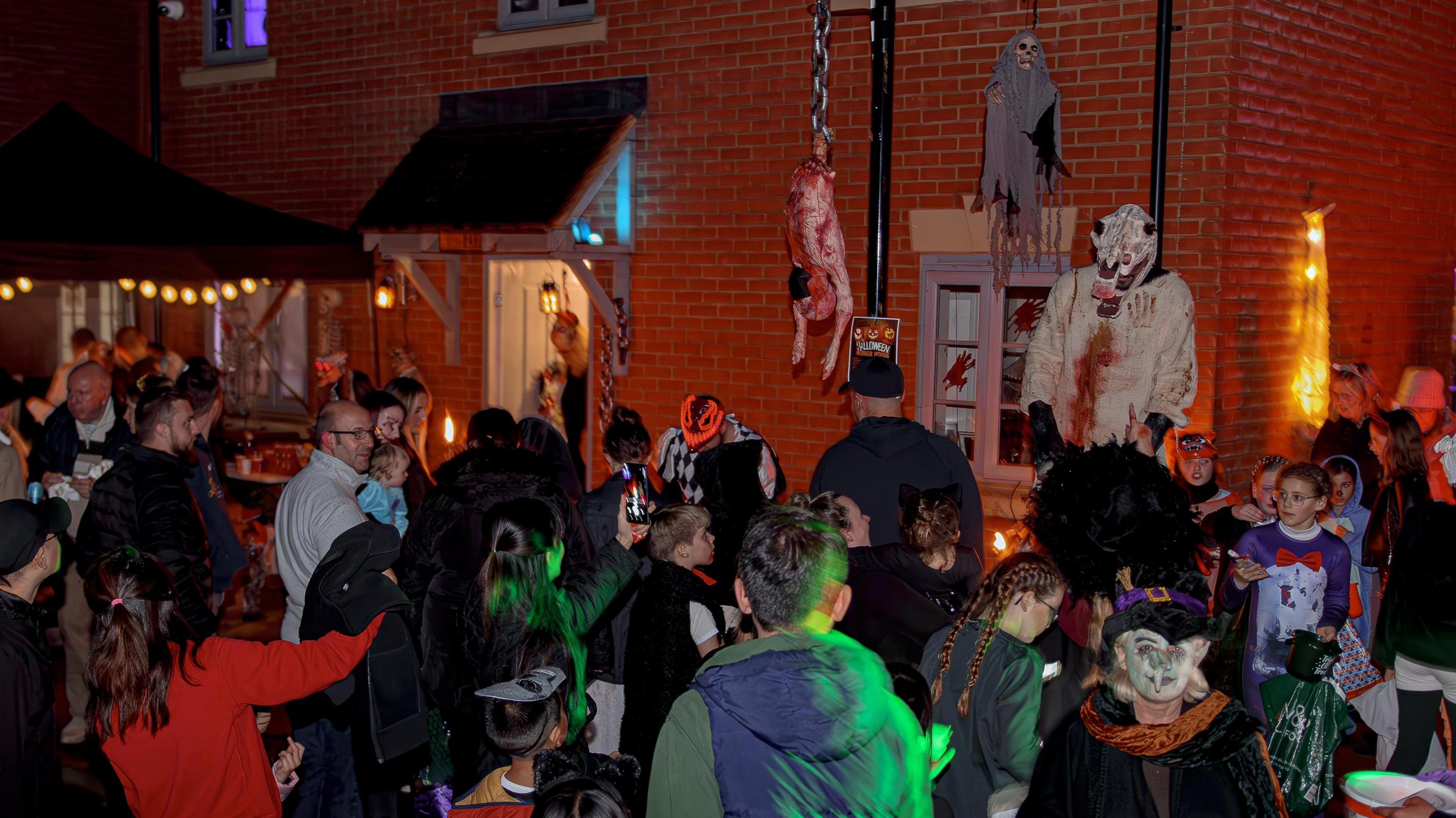 A large crowd of people dressed in various Halloween costumes. They are standing outside a red brick house covered in Halloween displays including bloody hand prints on the windows, a ghost in a grey shawl hanging from a wall, and a fake torso hanging from a chain.  