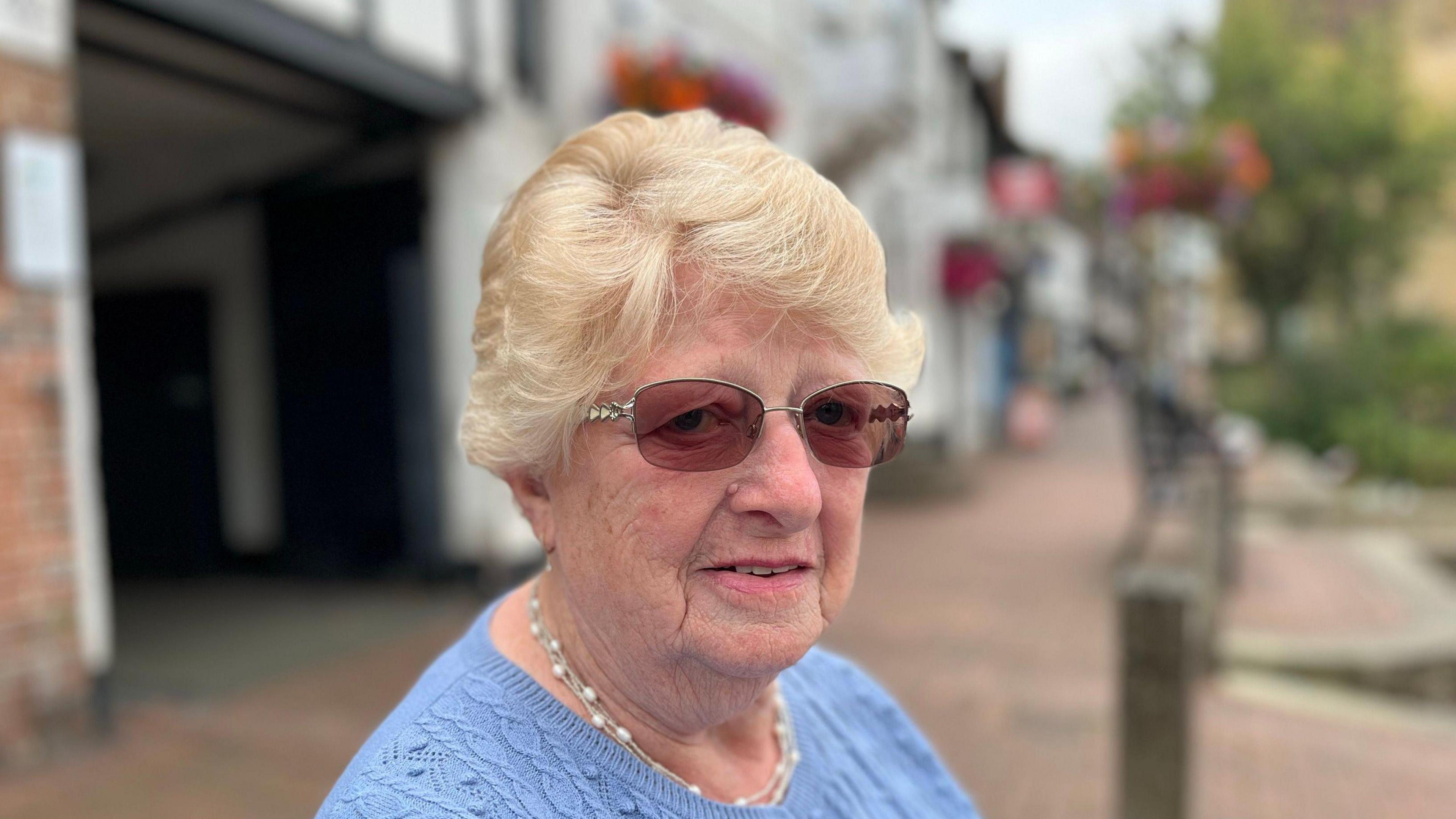 East Grinstead resident Shielah Harris pictured sitting on a bench in the high street wearing a blue cardigan, a pearl necklace and dark glasses