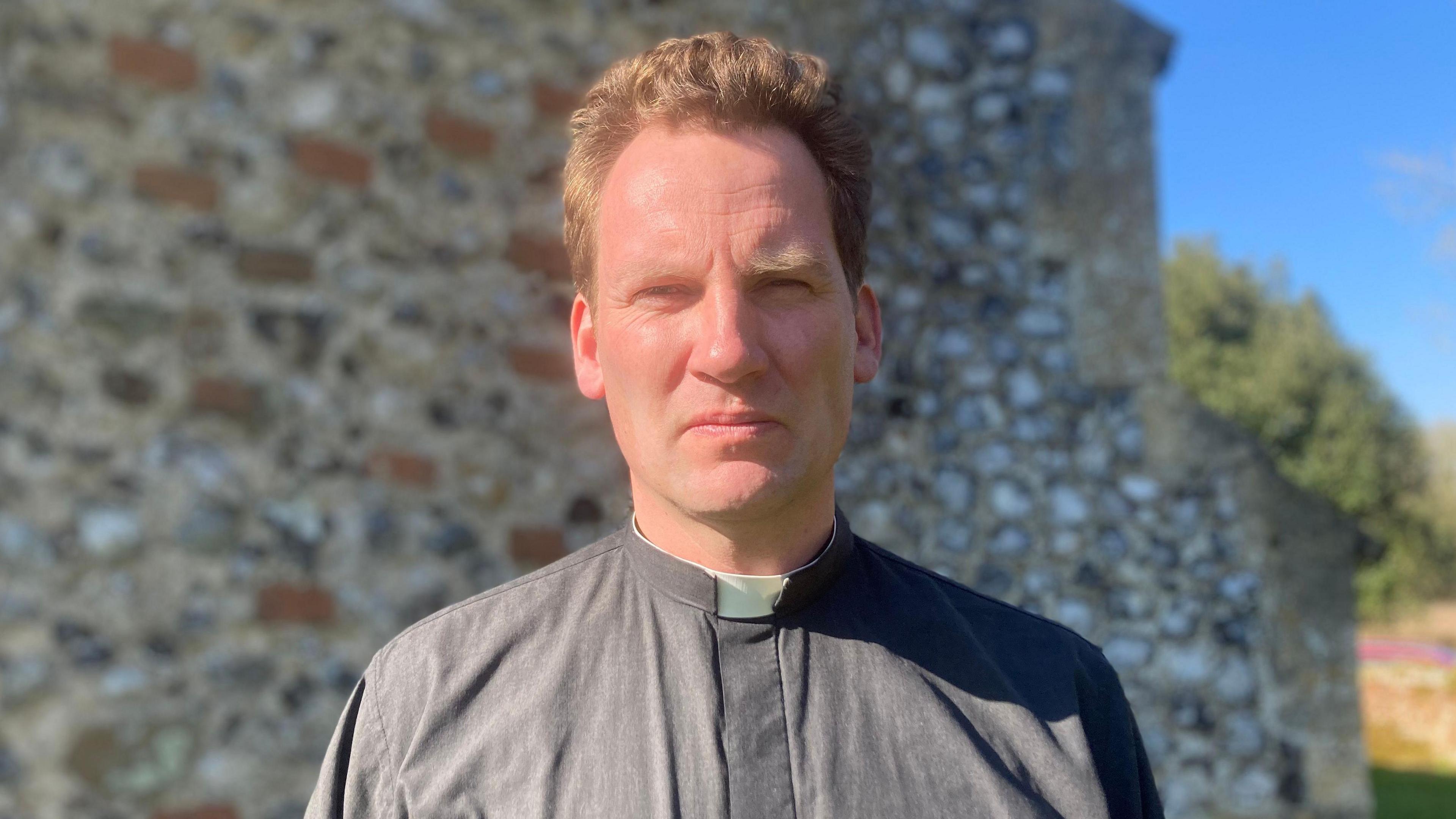 Reverend David Longe is standing in the middle of the picture. He has brown hair, and is smiling towards the camera. He is wearing grey vestment, with a white Church of England collar. He is standing in front of the flint walls of his church in Norfolk. 