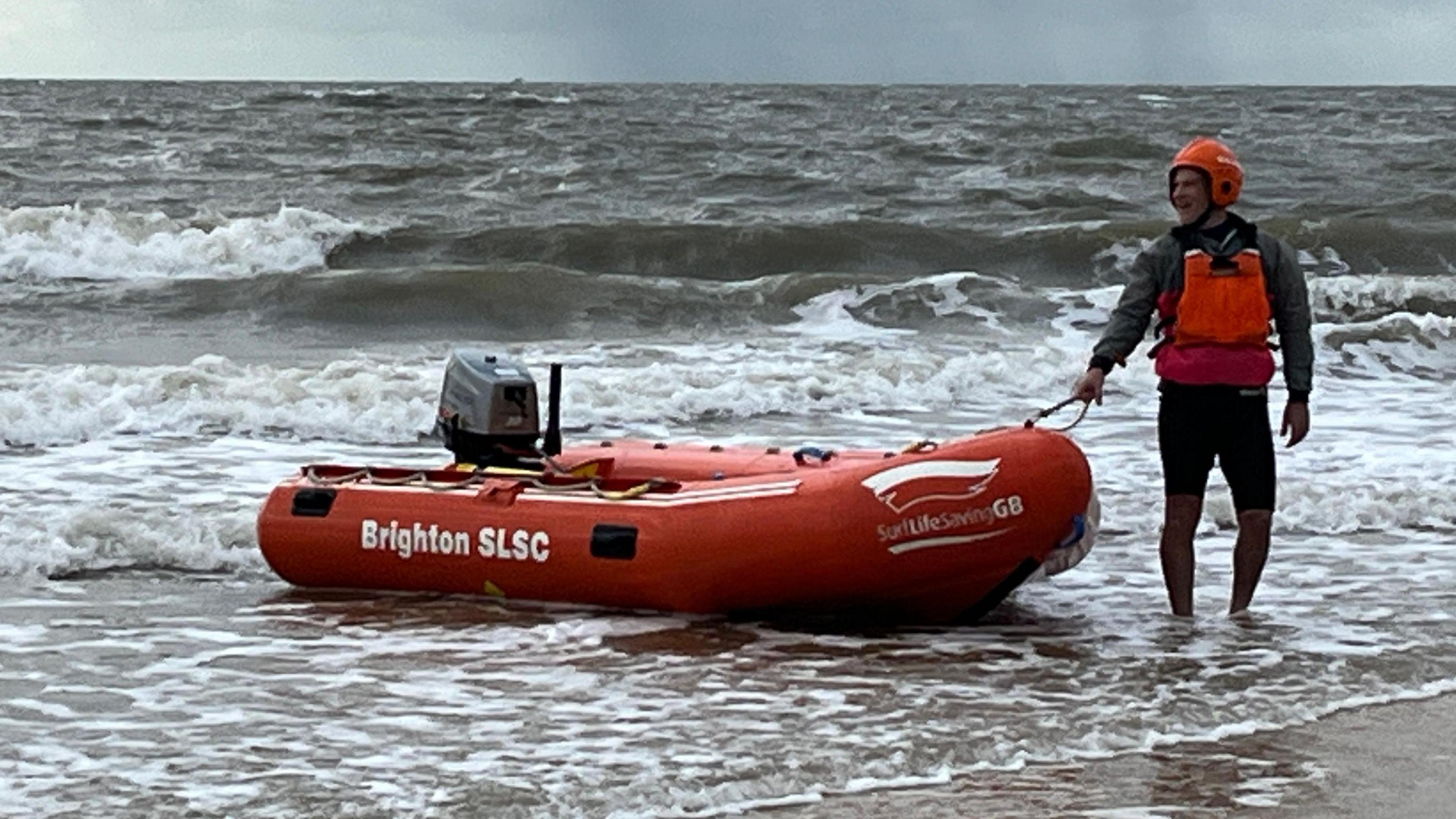 A man wearing a red helmet and red buoyancy aid holds a red dingy boat on the shoreline.