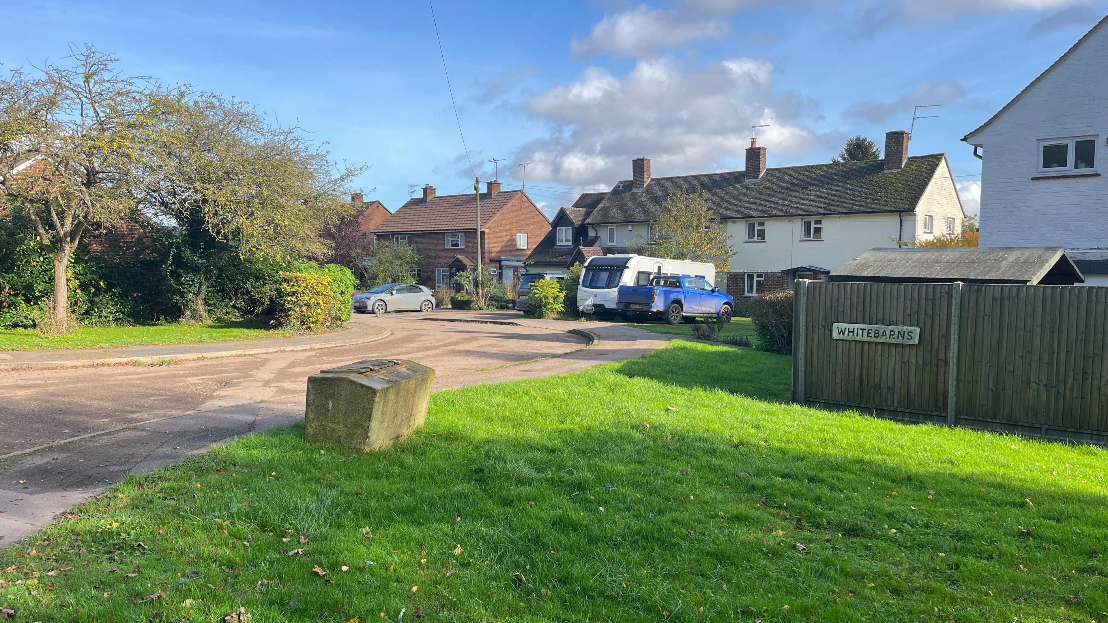 Several semi-detached local authority-style houses can be seen in a cul-de-sac. A car, pick-up truck and caravan can be seen parked outside. There is a blue sky with white and grey cloud.