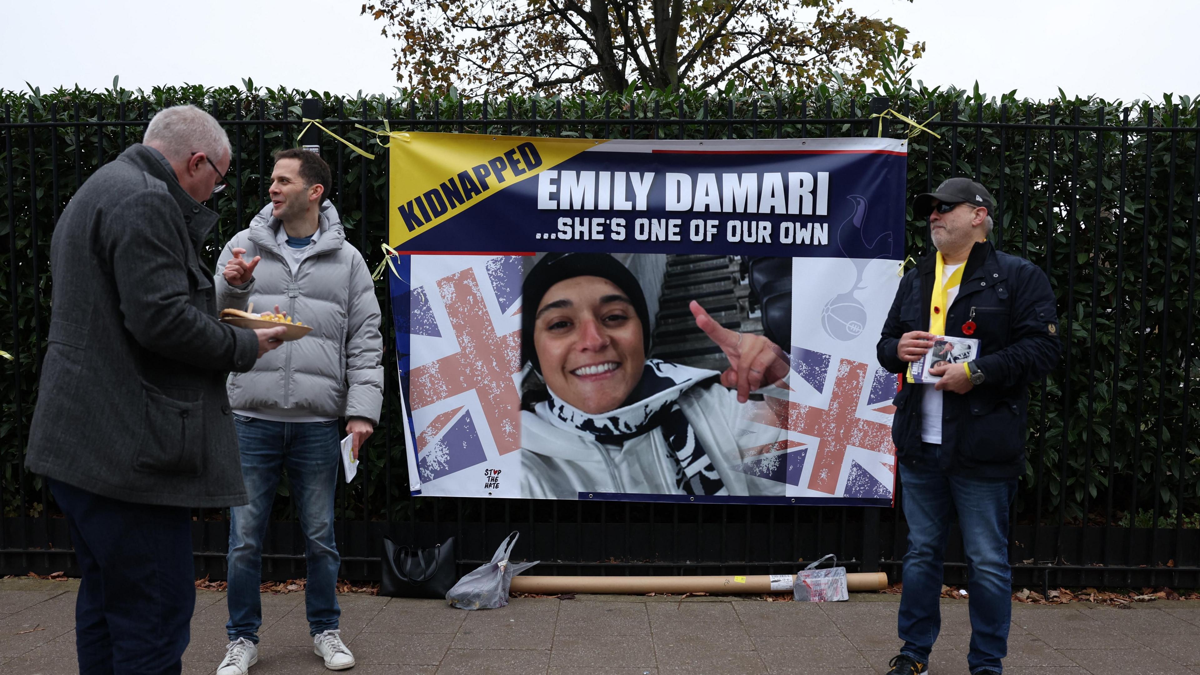 A large poster with a picture of Emily Damari, union jacks, and the word 'kidnapped' is hung on a hedge. Three men stand beside it, one with pamphlets in his hand.