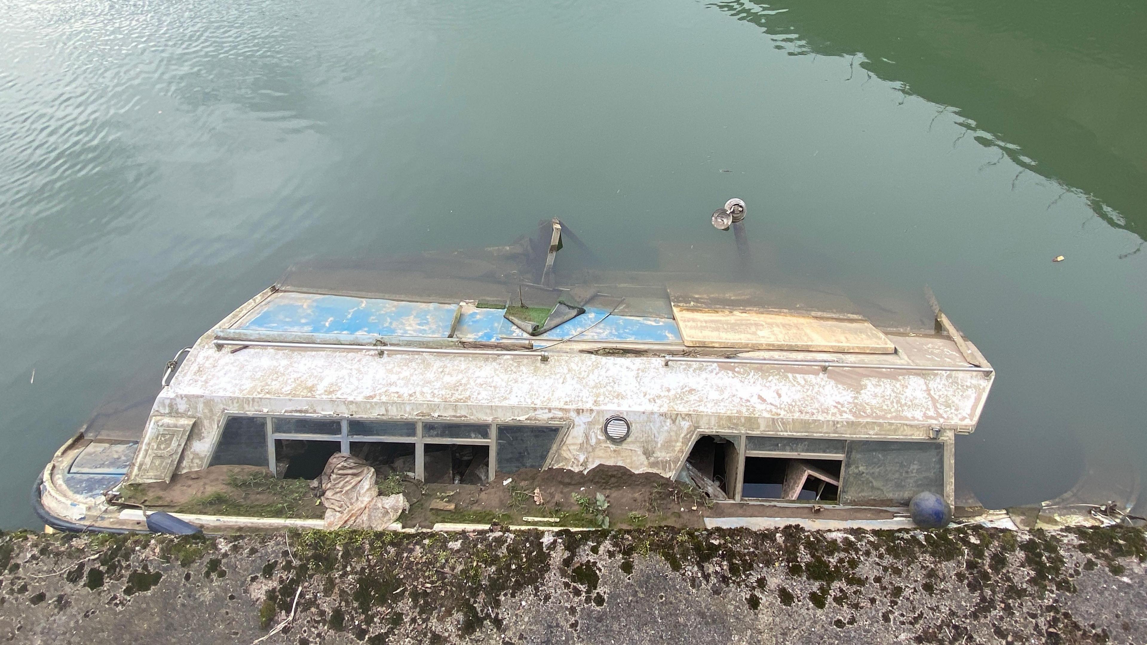 A boat partially submerged in the river in Bath. It is a large boat with debris to it's exterior and smashed windows.